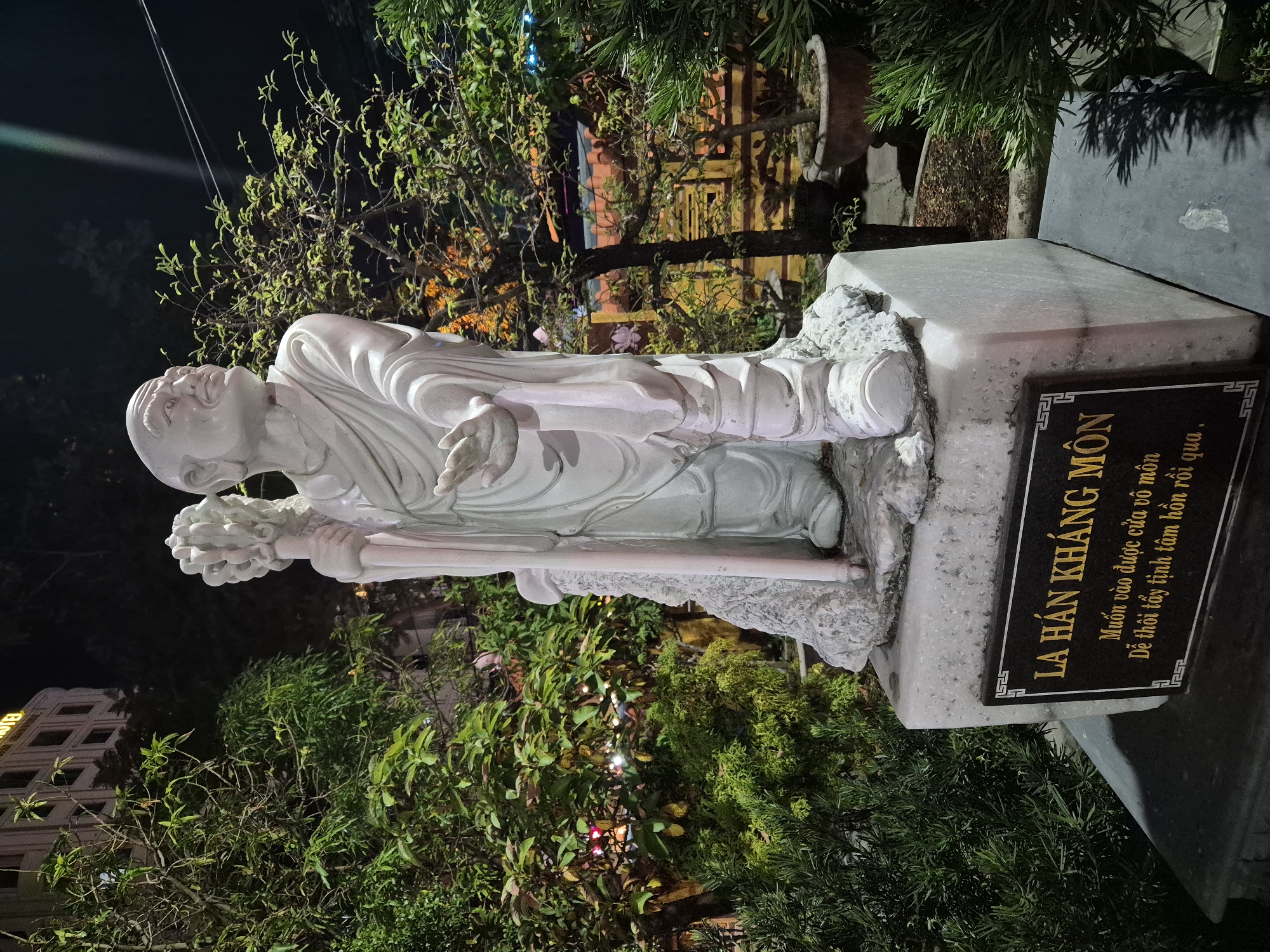 The image shows a white marble statue of a bald, smiling man in robes, holding a staff with a cluster of objects at the top. The statue stands on a rectangular stone base, which has a dark plaque with gold lettering. The plaque reads "LA HÁN KHÁNG MÔN" and below it, "Muốn vào được cửa vô môn / Dễ thôi tẩy tịnh tâm hồn rồi qua." The statue is surrounded by lush green trees and bushes, with a building visible in the background. The lighting suggests it is nighttime, with artificial lights illuminating the scene and creating shadows. The building in the background appears to be a hotel, indicated by the word "HOTEL" in large lettering. Based on the text on the plaque, which is in Vietnamese and likely refers to Buddhist concepts, and the architectural style of the building, the location is likely a temple or a public space in Vietnam, possibly in the city of Đà Nẵng.