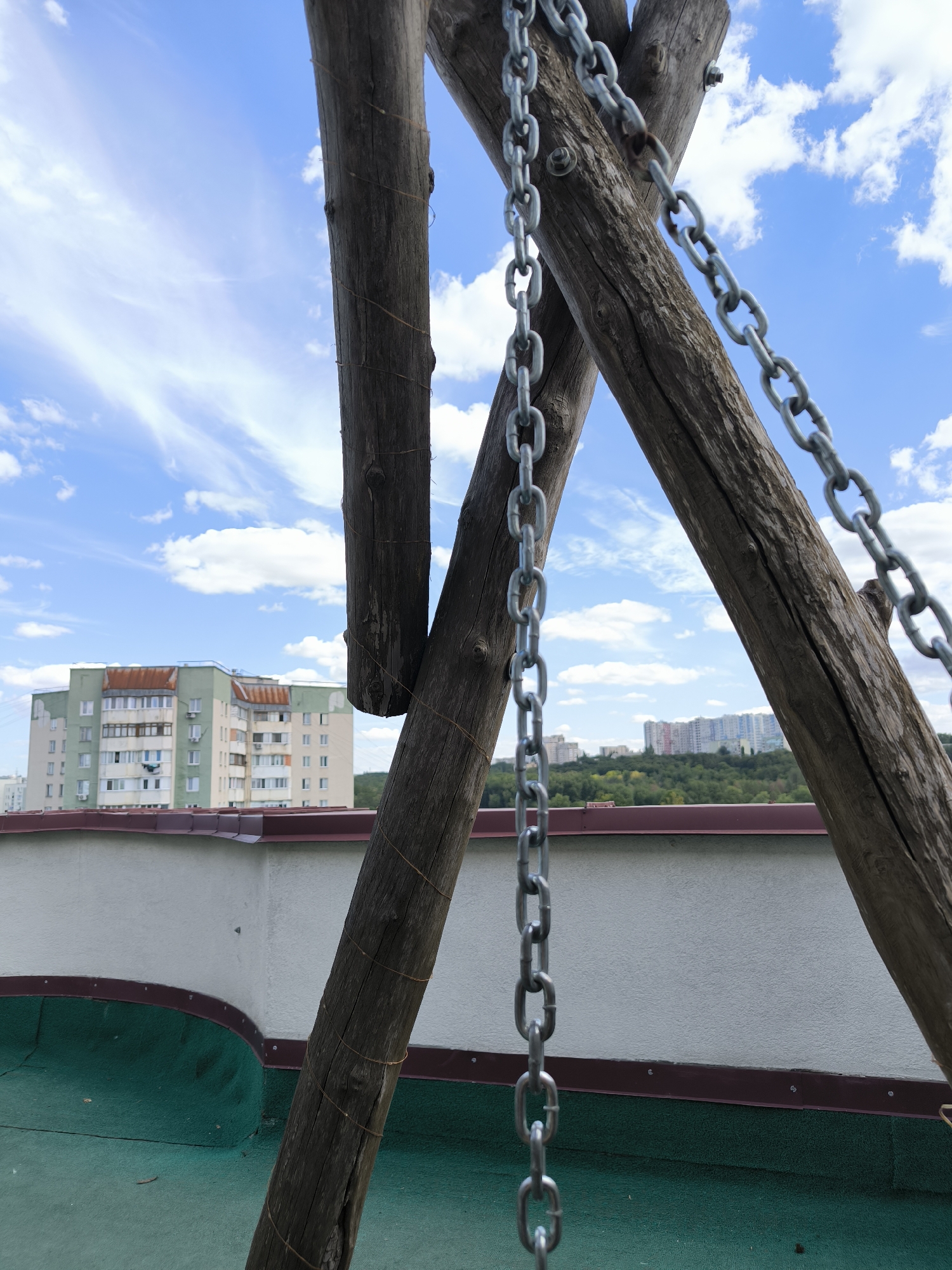 The image captures a close-up view of a wooden swing set structure with metal chains hanging from it, set against a backdrop of a bright blue sky with scattered white clouds. In the middle ground, there are residential buildings, a green forest, and a rooftop with a green surface and white walls. The scene appears to be from a rooftop in the city of Chișinău, Moldova, likely in a residential area. The wooden beams are weathered and show signs of age, with thin wires wrapped around them. The chains are metallic and appear to be part of a swing set. The buildings in the background are multi-story and have a mix of architectural styles. The overall impression is one of an outdoor recreational area on a clear day. There are no people or animals visible in the image.