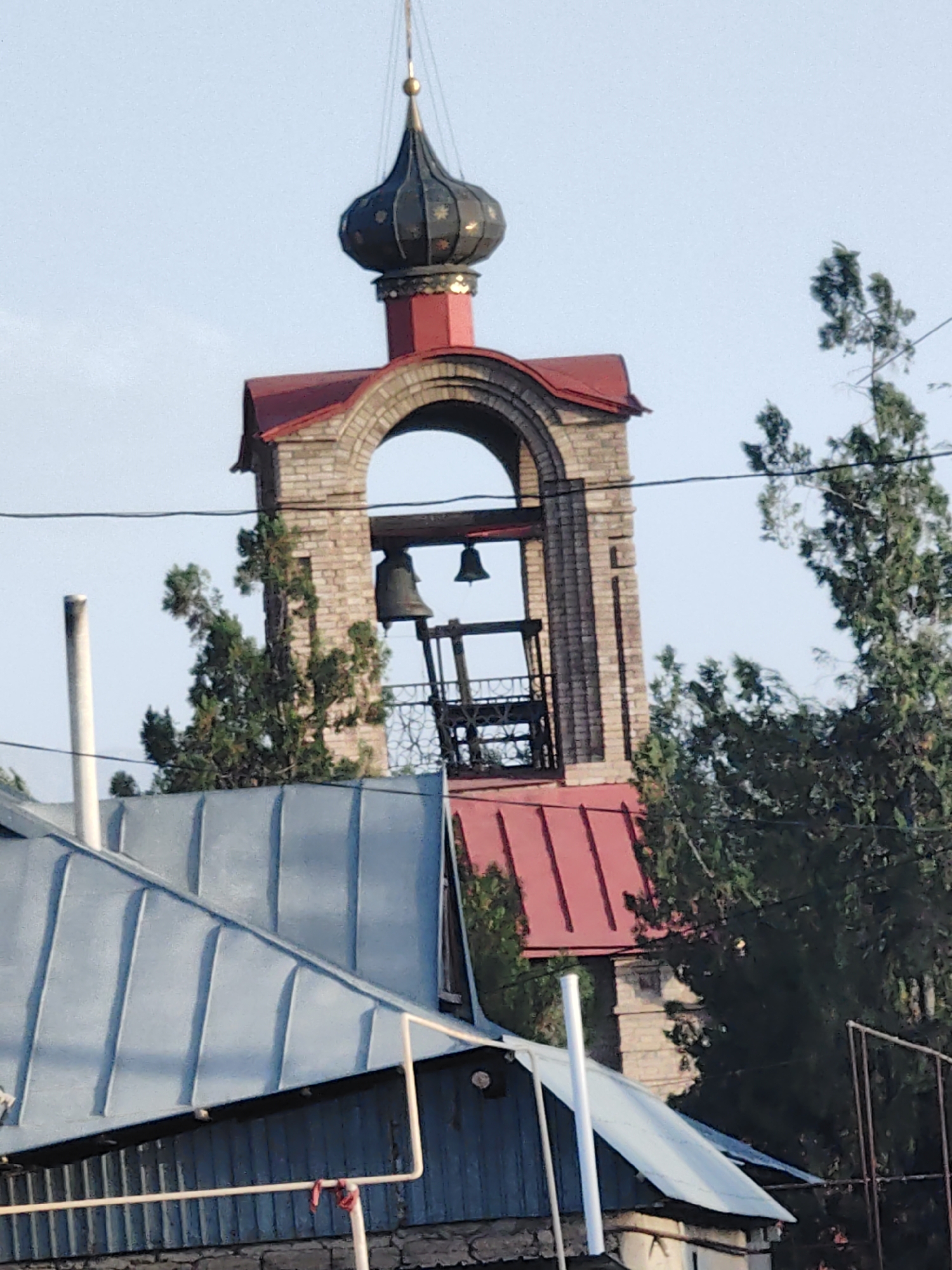 The image shows a close-up, eye-level, outdoor, daylight, medium shot of a church bell tower. The bell tower has a red roof and is made of brick. There are two bells hanging in the tower. In the foreground, there is a blue metal roof with a white pipe running along it. In the background, there are green trees and a clear blue sky. The location is in the village of Koksayaq, in the Koksayaq rural district of Kazakhstan.