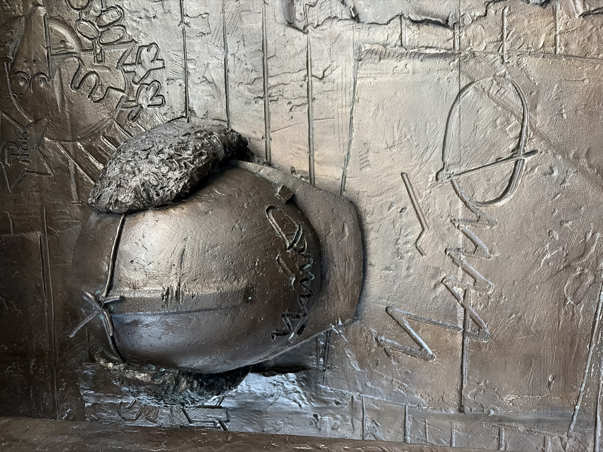 The image is a close-up view of a bronze relief sculpture, likely part of a larger monument. The sculpture depicts a stylized head wearing a cap with fuzzy ear flaps. The face is obscured by shadow and the bronze material. Below the head, a signature or inscription is carved into the bronze surface, appearing as a series of angular lines and a circular shape with a line through it. The background of the relief features textured lines and what appear to be abstract or symbolic elements, such as a star shape with lettering above it, and a snowflake-like pattern. The sculpture has a metallic, aged patina. This artwork is located on the facade of the Yekaterinburg State Academic Opera and Ballet Theatre in Yekaterinburg, Russia, and is part of a bas-relief dedicated to the history of the theatre.