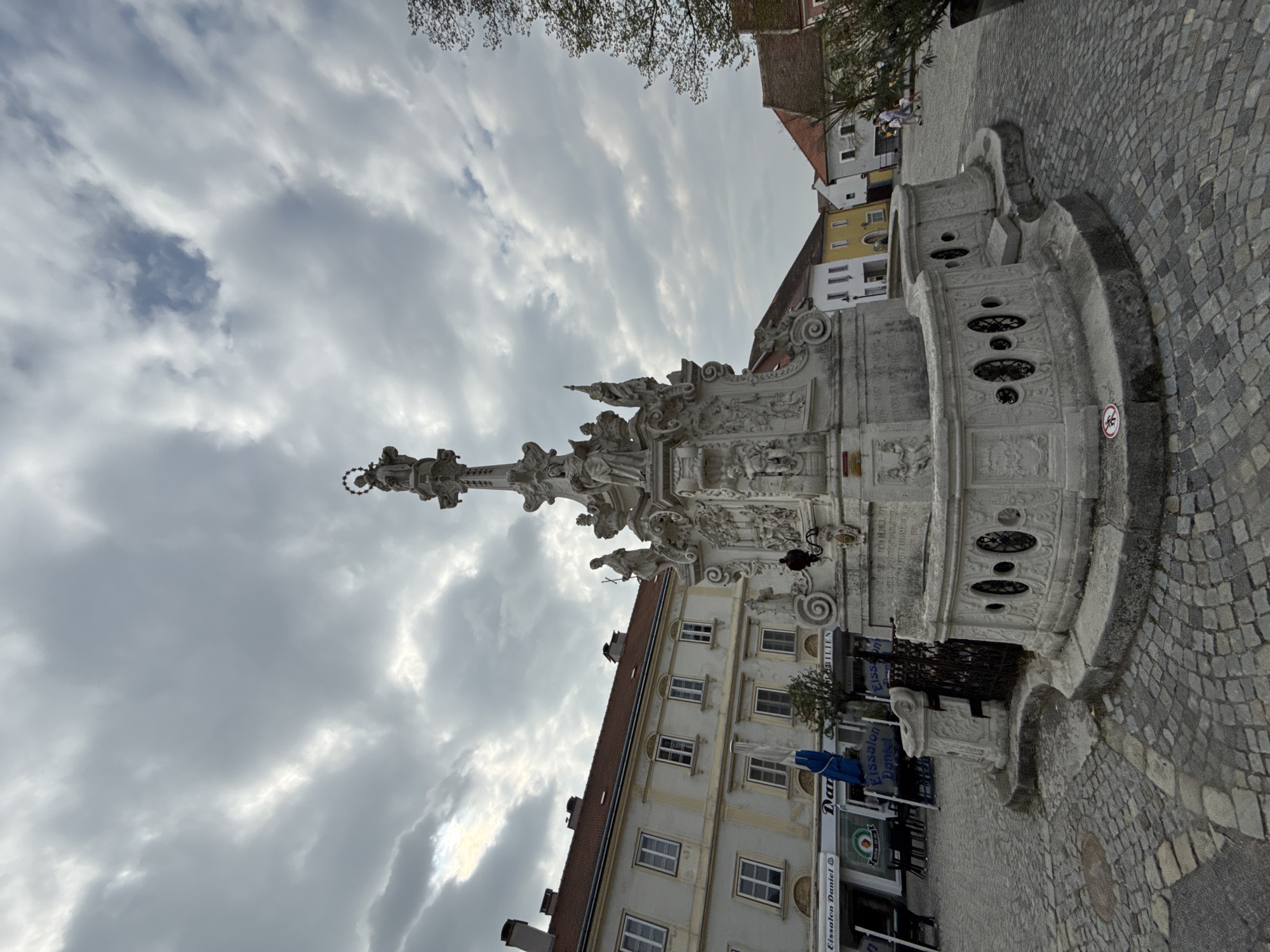 The image displays a grand, ornate white stone monument situated in a cobblestone town square in Hainburg an der Donau, Austria. The monument, centrally positioned in the scene (when correctly oriented), is intricately carved with numerous Baroque-style sculptures, including statues of figures, decorative reliefs, and a tall central column topped with a crowning figure. Its wide base features several tiers with arched openings and stone railings.
The ground around the monument is paved with textured gray cobblestones. Surrounding the square are traditional European buildings, some with multiple stories. A prominent building in the background features a reddish-brown tiled roof and a light-colored, possibly yellow or off-white, facade with numerous rectangular windows. Other buildings and hints of shopfronts are also visible. Sparse tree branches are seen against the sky at the top of the frame.
The sky is heavily overcast with large, diffused clouds, indicating a daytime scene. While the weather is cloudy, brighter patches suggest light attempting to penetrate the cloud cover. A few indistinct human figures can be seen near the buildings in the background, but no specific activity is clearly taking place.
Visible text includes "Eissalon Daniel" on a shop sign, another sign partially reading "Dan...", and "immobilien" on a building facade. A red circular prohibitory sign is also visible on the cobblestones near the base of the monument.