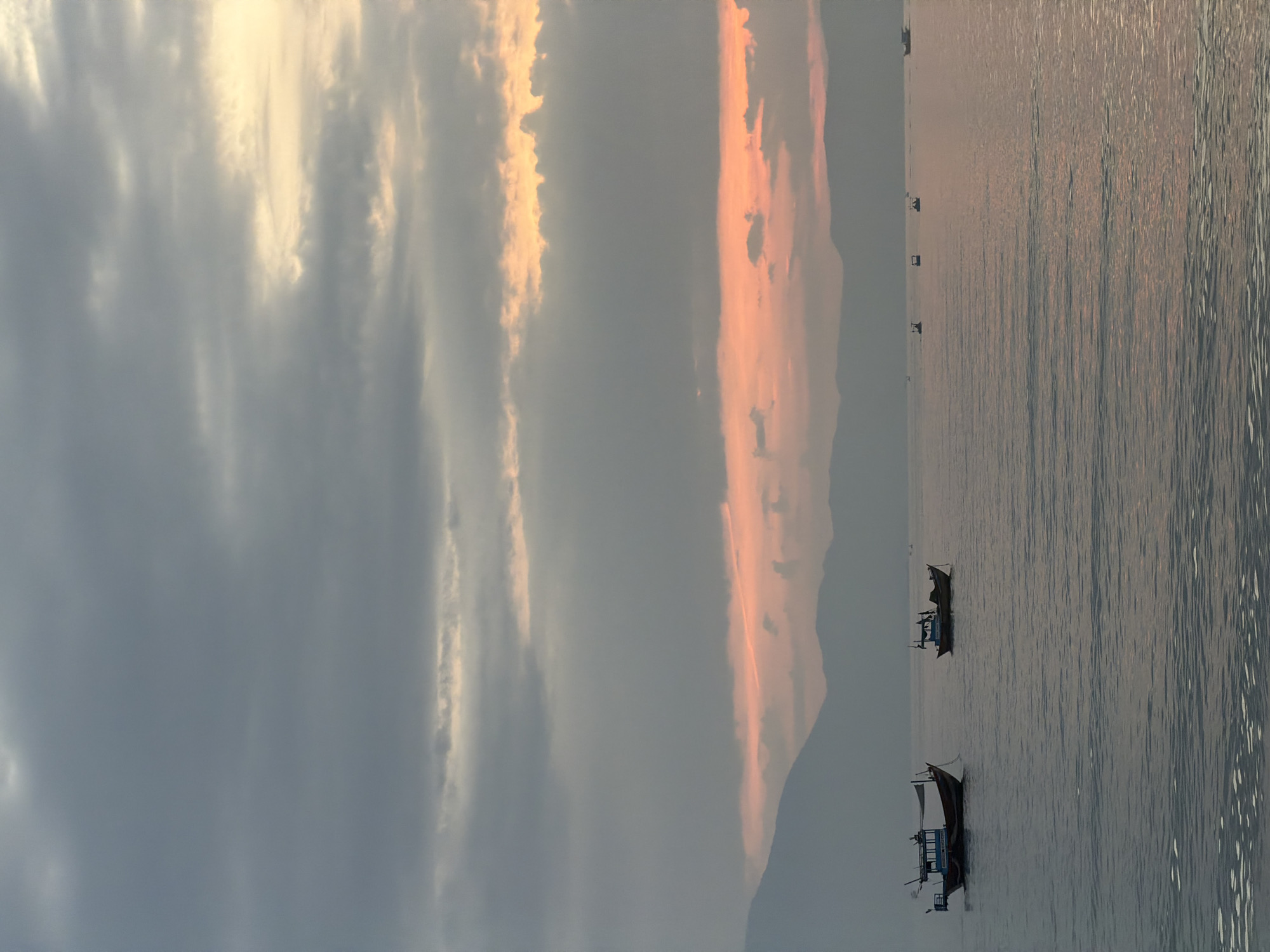 At dusk, two small fishing boats are anchored on a calm sea. The boat in the foreground to the left is larger than the boat on the right. The boats are silhouetted against the water, which reflects the muted colors of the sky. In the background, a range of hills is visible, also silhouetted against the horizon. The sky is filled with clouds, with streaks of orange and pink indicating the setting sun. The overall scene is peaceful and serene, characteristic of a quiet evening on the coast of Dong Ninh Hoa Ward, Vietnam.