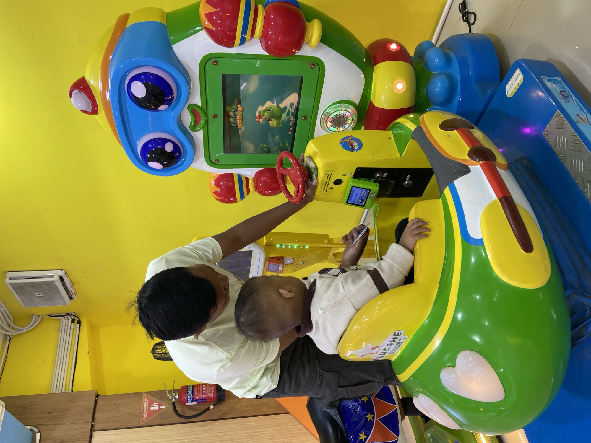 A father is helping his young child sit in a colorful children's arcade game in an amusement center. The child is seated in a green and yellow car-shaped game with a red steering wheel. The father, wearing a light-colored t-shirt and dark pants, is looking down at the child. The game features a screen displaying a cartoon character, likely part of a game. The walls of the amusement center are painted bright yellow. A fire extinguisher is visible on the left side of the image. The game itself is large and resembles a robot. The floor appears to be tiled. The overall setting suggests a fun, family-friendly environment within Kecamatan Pati, Indonesia.