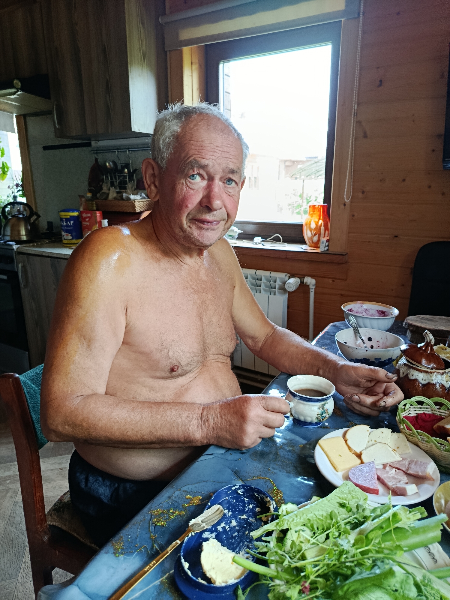 An elderly Caucasian man with gray hair and blue eyes is sitting at a table indoors, holding a cup of coffee. He is shirtless and appears to have been sweating, with a sheen on his shoulders. The table is covered with a blue tablecloth and set with various food items, including a plate of sliced cheese and ham, a bowl of what appears to be berries and cream, a basket with bread, and a dish with butter and a spoon. A kettle and other kitchen items are visible in the background. The room has wooden walls and a window with a view of the outdoors. The man is looking directly at the camera with a neutral expression.