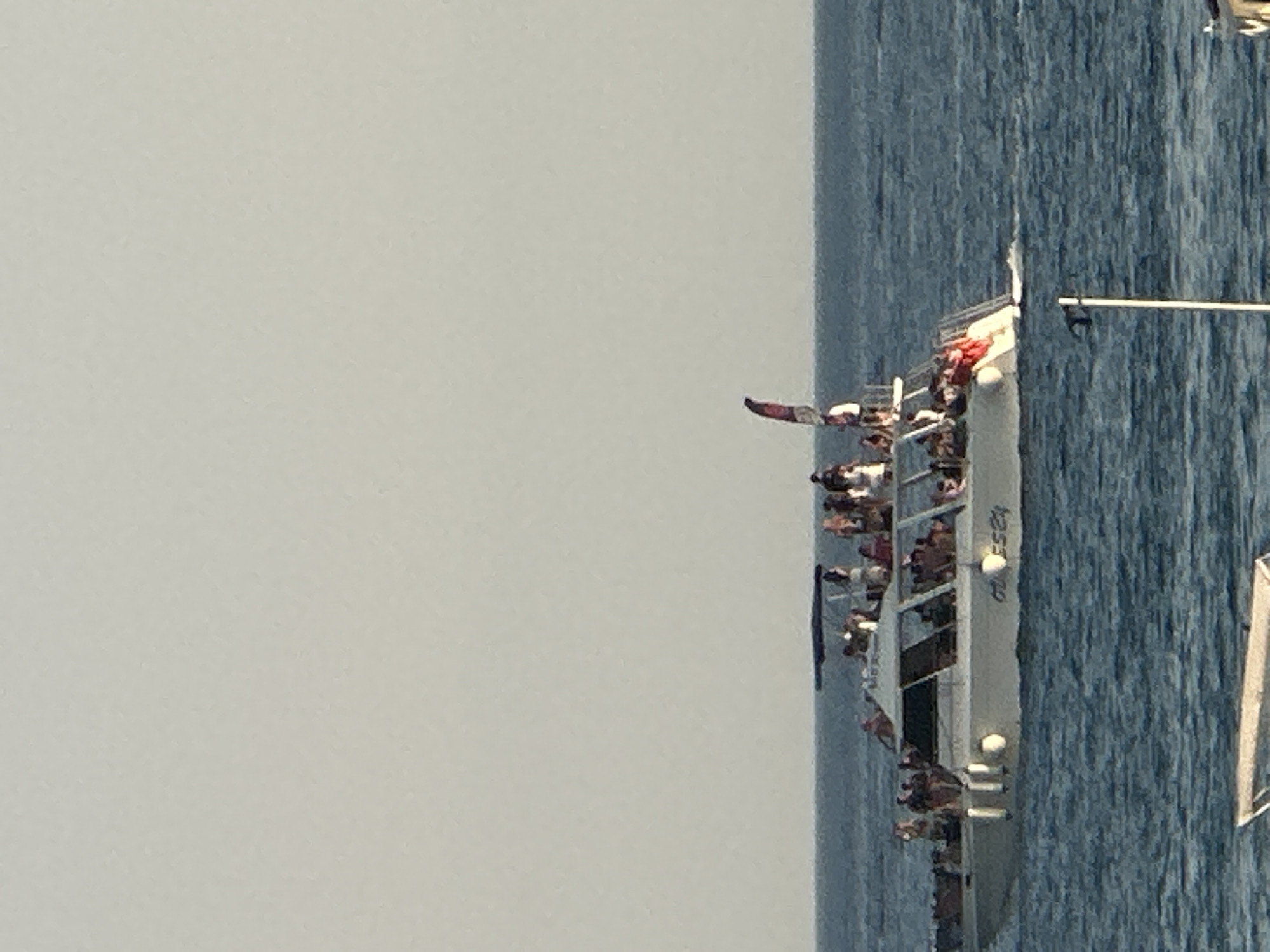 The image shows a boat filled with people in the water near Boreti, Montenegro. The boat is a white catamaran with a dark canopy. People are standing and sitting on the upper deck and in the enclosed cabin. Some people appear to be wearing red clothing. A flag with red and white colors is visible on the boat. The water is a deep blue, and the sky is a pale gray. In the foreground, a small part of another boat is visible. The overall scene suggests a sunny day with people enjoying a boat trip.