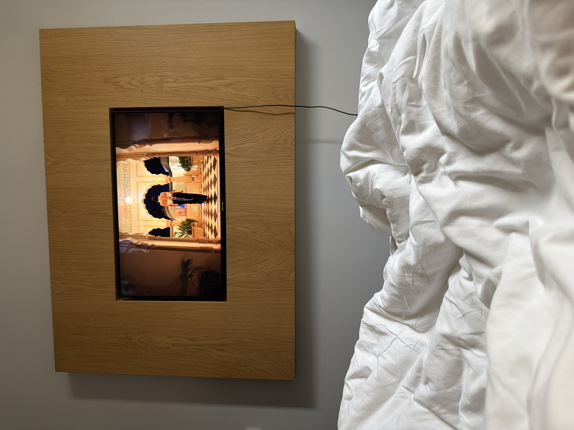 A wooden-framed television screen is mounted on a grey wall, displaying an image of what appears to be an interior scene of a building in Tbilisi, Georgia, featuring arched doorways and decorative elements. In the displayed scene, two individuals are visible, one wearing a black suit. A black cable extends from the frame of the television towards a crumpled white bed sheet that partially obscures the right side of the frame.