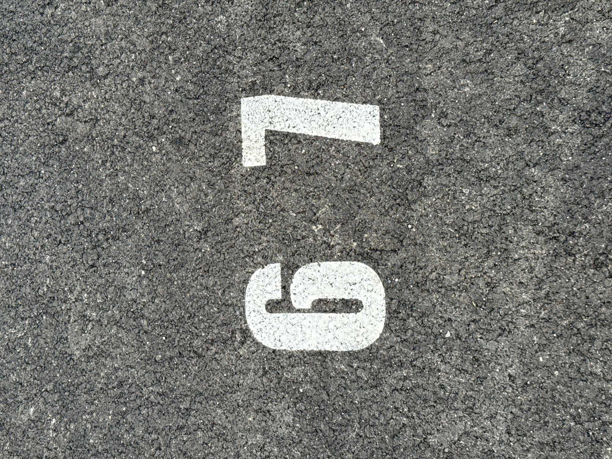 The image is a top-down view of a dark asphalt surface with two large white numbers, "6" and "7", stenciled on it, representing the number "67". The asphalt has a textured, granular appearance with small lighter colored particles. The lighting suggests it is outdoors in daylight. The location of this image is unknown, but if it were in Burgas, Bulgaria, it could potentially be a parking space number, a marker on a sports field, or part of an industrial marking.