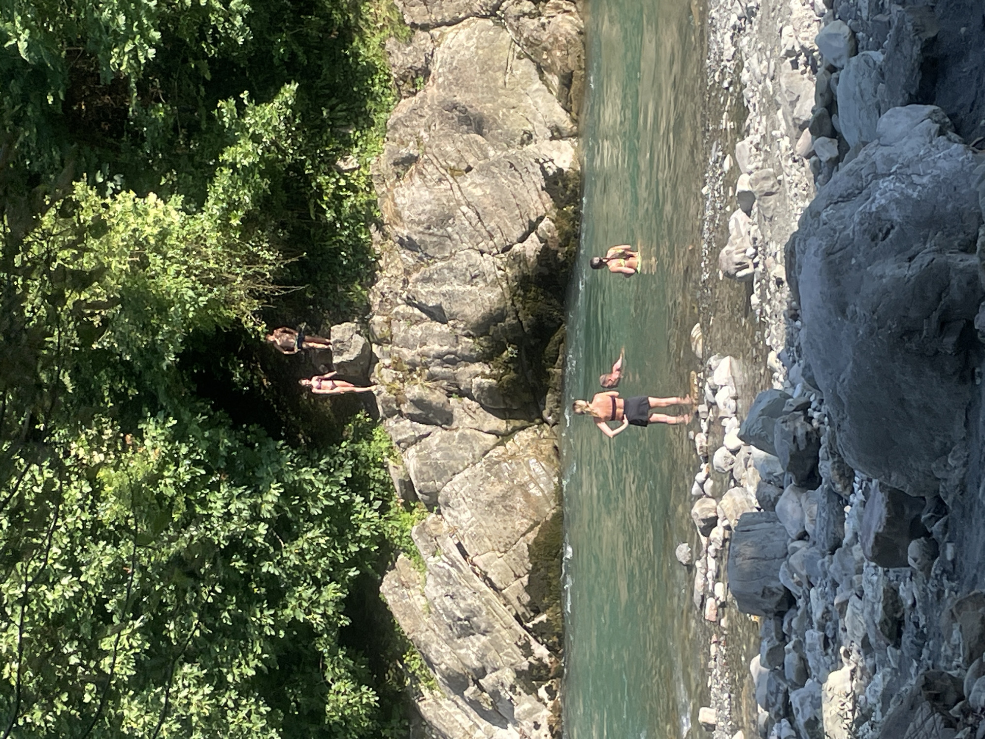In Sochi, Russia, on a sunny day, several individuals are enjoying a clear, turquoise river. The river flows over a rocky bed, with large boulders scattered along the shoreline and within the water. Lush green trees and dense foliage form a canopy over the scene, casting shadows on the rocky cliffs that rise behind the river.
On the bank, on a rocky ledge, three people are visible, two women and one man. One woman, wearing a bikini, stands on a rock. Another woman in a bikini is near the water's edge. A man in shorts stands on a rock.
In the river, one woman stands waist-deep in the water, wearing a black skirt and a life vest. Further out, another woman in a yellow bikini is also in the water. A third person is partially submerged, their head visible. The water is calm, with gentle ripples indicating a slow current. The overall impression is one of a serene and natural setting, ideal for recreation.