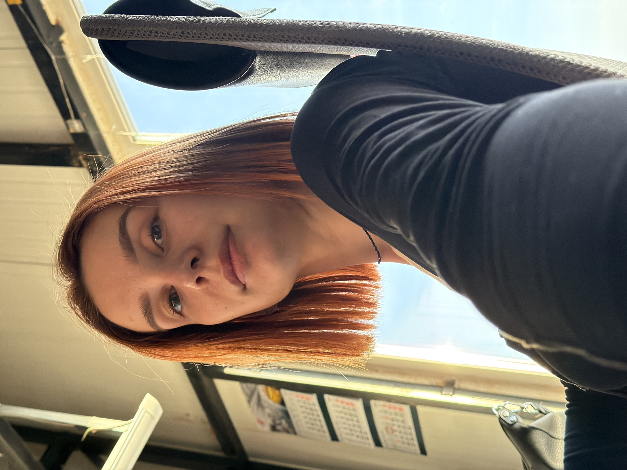 A reddish-haired young woman is visible, tilting her head downward toward the camera. She wears a dark long-sleeved shirt. A black chair or headrest is positioned above her. A window frame and a glimpse of the sky are visible in the background. There is also a calendar on a wall. The location is specified as Chaikovsky, Russia. No specific actions or interactions are evident.