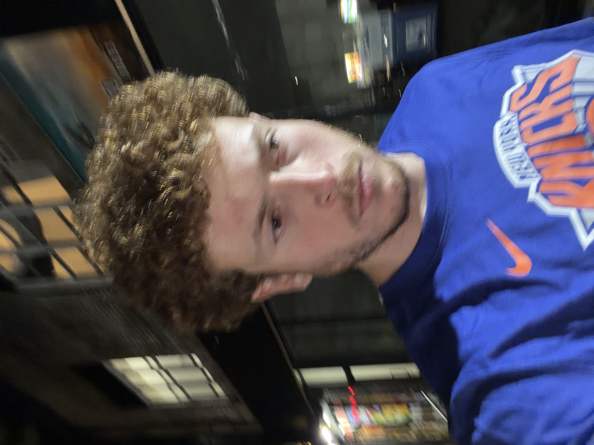 A photograph depicts a person with light skin, light brown curly hair, and a short beard, wearing a blue sweatshirt with "New York Knicks" printed on it, along with a visible orange Nike swoosh logo. The background appears to be an urban setting at night, with blurred lights and building structures visible. The image seems to be taken in a moving or unsteady manner, as indicated by the blur. Specific details suggesting the location being the city of New York are the New York Knicks apparel and the urban backdrop.