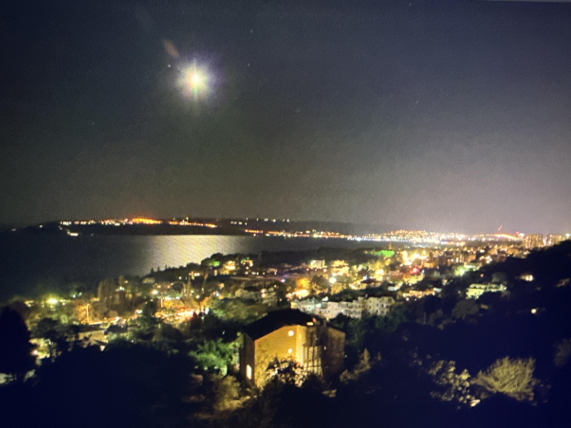The image shows a nighttime view of a coastal city. A body of water is visible on the left, with a stretch of land in the distance illuminated by lights. The city has scattered building lights and streetlights. In the foreground, there are dark, silhouetted trees and vegetation, with a single building illuminated by a yellow glow, standing out amongst the darker landscape. Above, the sky is dark with a bright point of light, possibly a celestial body. No people are visible in the image. The location cannot be confirmed as Sofia, Bulgaria.