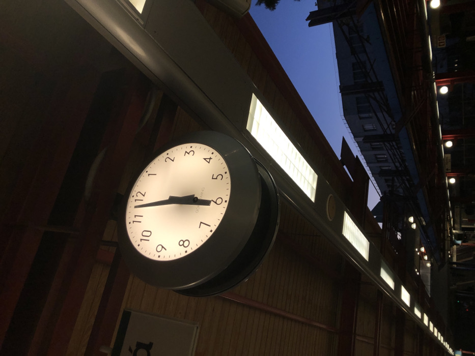 The image captures a tilted, upward-looking view of an indoor or semi-enclosed public space, likely a train or metro station in Moschato, Greece, during the evening or night. The main subject is a large, round, illuminated analog clock, prominently displaying the time as approximately 5:45. The clock has a white face with black numbers and hands, and the text "MING" is partially visible below the center.
The clock is suspended from an overhead structure, which extends across the frame and features a series of rectangular, brightly lit artificial light panels. These lights contrast with the dark blue sky visible in the upper right, where the silhouettes of distant buildings can be seen, confirming the time of day as dusk or night.
Below the clock, a structured wall or ceiling section with dark, vertical panelling is visible. A white sign with the Greek letter "ά" is partially seen in the lower left quadrant. The scene is devoid of people, focusing instead on the architectural elements and the functional clock, which serves as a central point of interest in the artificially lit environment.