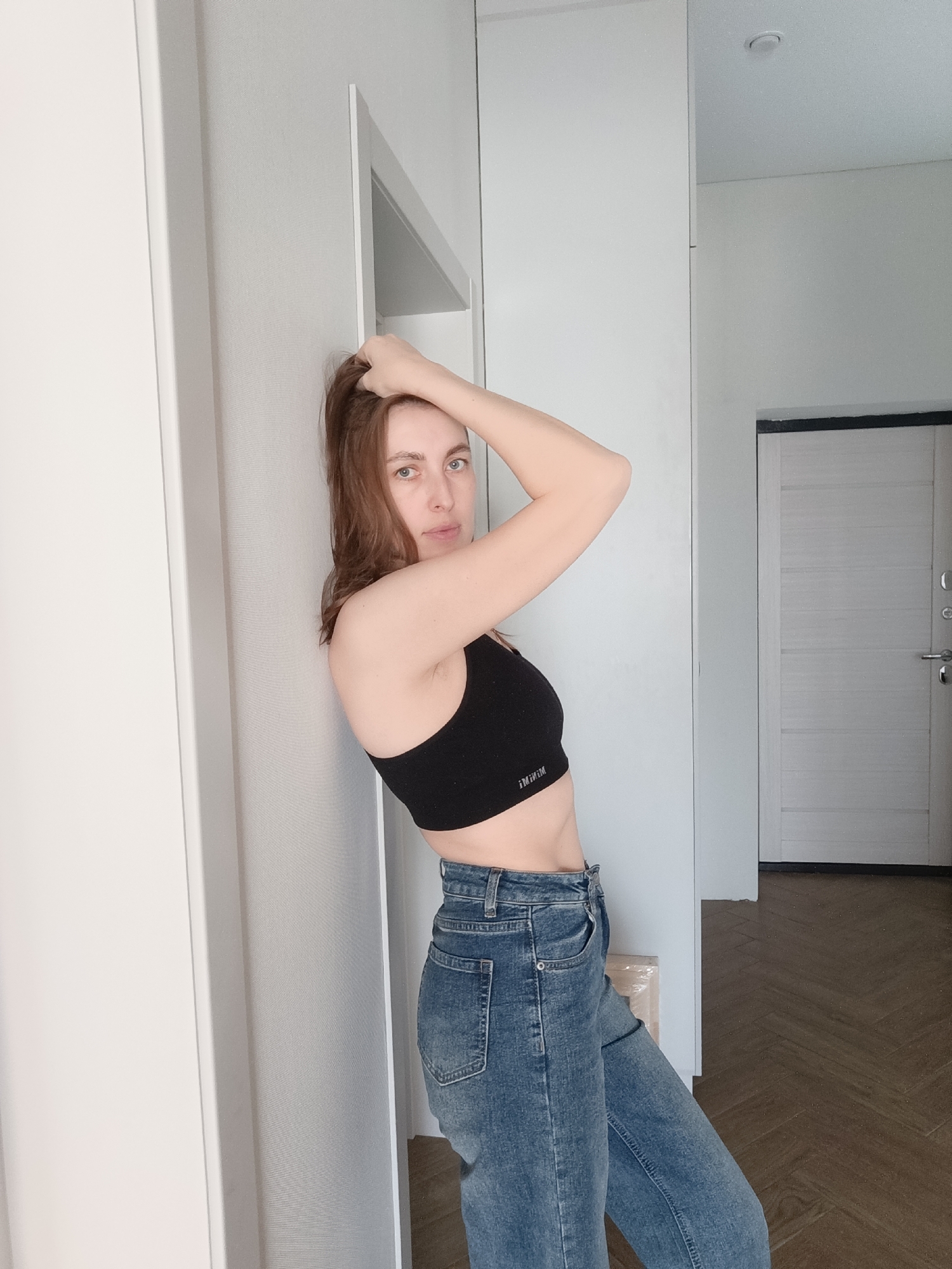 A woman is standing in an interior hallway or room. She is leaning against a white wall, facing the camera. The woman has shoulder-length brown hair that she is touching with her right hand. She is wearing a black sports bra and high-waisted blue jeans. The floor is a herringbone pattern in dark brown and light brown tones. In the background, there is a white door with a gray frame. The location is Chaikovsky, Russia.