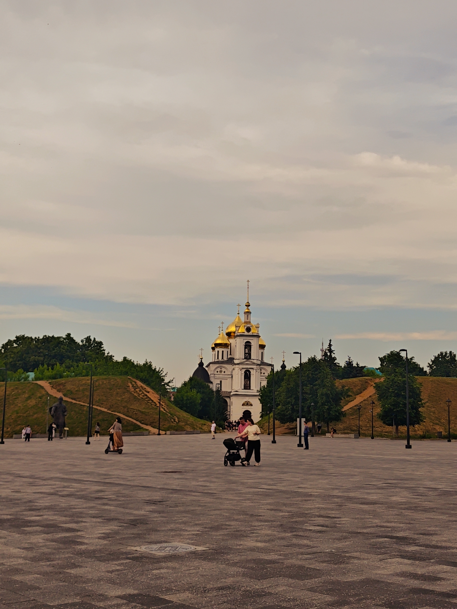 In the city of Unknown, Unknown, an outdoor plaza features a large, paved area with a textured pattern of grey squares. A white cathedral with golden domes is visible in the background, nestled among lush green trees. To the left of the cathedral, a grassy hill rises, topped with trees. A large, dark-colored statue stands on the left side of the hill. Several people are scattered across the plaza: a woman rides a scooter, two women push a stroller, and other individuals walk or stand in small groups. Lampposts with black poles are positioned throughout the plaza. The sky above is a pale, overcast grey.
