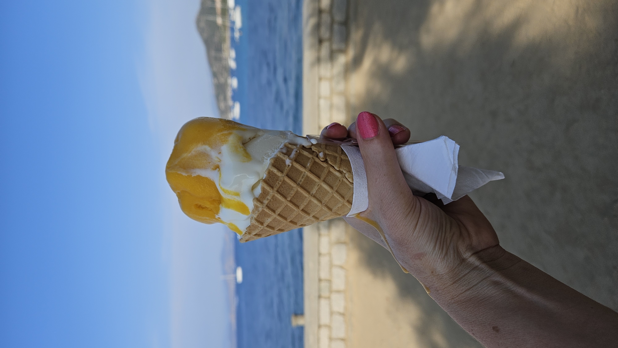 A hand with bright pink nail polish holds a melting ice cream cone with two scoops of ice cream, one white and one orange, against a backdrop of a bright blue sky and the ocean. In the background, several white sailboats are anchored in the water, and a hilly coastline with trees is visible in the distance. A low stone wall runs horizontally across the mid-ground. The scene is outdoors during the daytime, suggesting a sunny, coastal environment. While the specific location within L'Île-Rousse, France, is not identifiable from the image alone, the presence of sailboats and a Mediterranean-style coastline is consistent with the area.