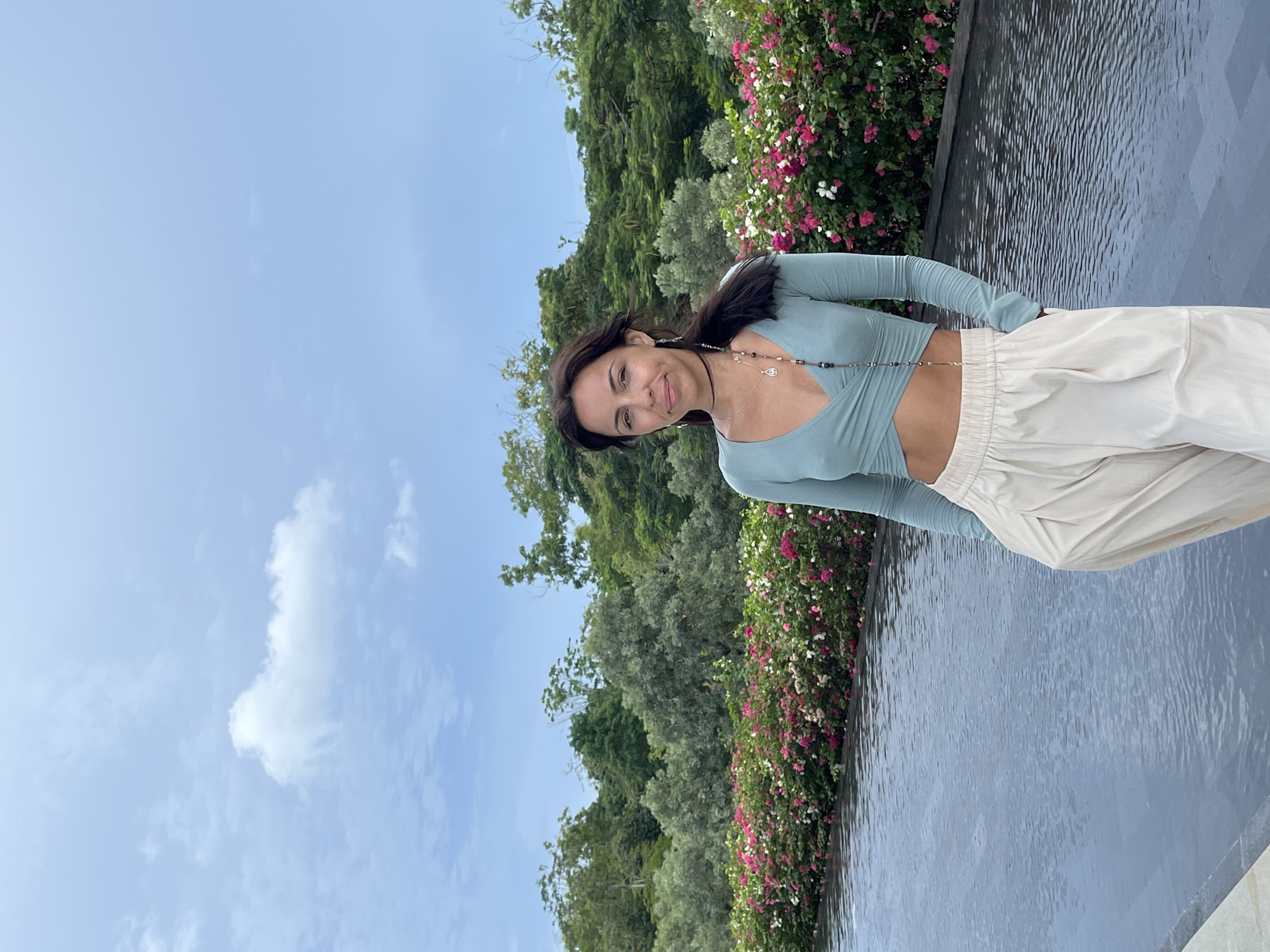 The original image is presented rotated 90 degrees counter-clockwise. When viewed correctly oriented, it captures a young woman posing outdoors on a bright day in Kuta Selatan, Indonesia.
The woman stands on the right side of the frame, facing the camera with a gentle, composed smile. She has dark hair, pulled back slightly, and a tanned complexion. She is wearing a light teal, long-sleeved crop top with a V-neck and a wrap-around design, revealing a portion of her midriff. Her lower body is clad in loose-fitting, high-waisted, off-white or beige pants with an elastic waistband. She accessorizes with several delicate necklaces, including a beaded choker and a longer chain that appears to drape across her torso, possibly a waist chain, along with visible earrings.
The background on the left features lush green trees and varied foliage under a clear, light blue sky with a few wispy white clouds. On the right, behind the woman, is a vibrant bush adorned with numerous pink and white flowers, likely bougainvillea. Below this flowering bush, a tranquil body of water, possibly a decorative pool or pond, stretches across the lower right portion of the frame, its surface reflecting light with subtle ripples. The scene evokes a sense of calm and natural beauty. No visible text is present in the image.