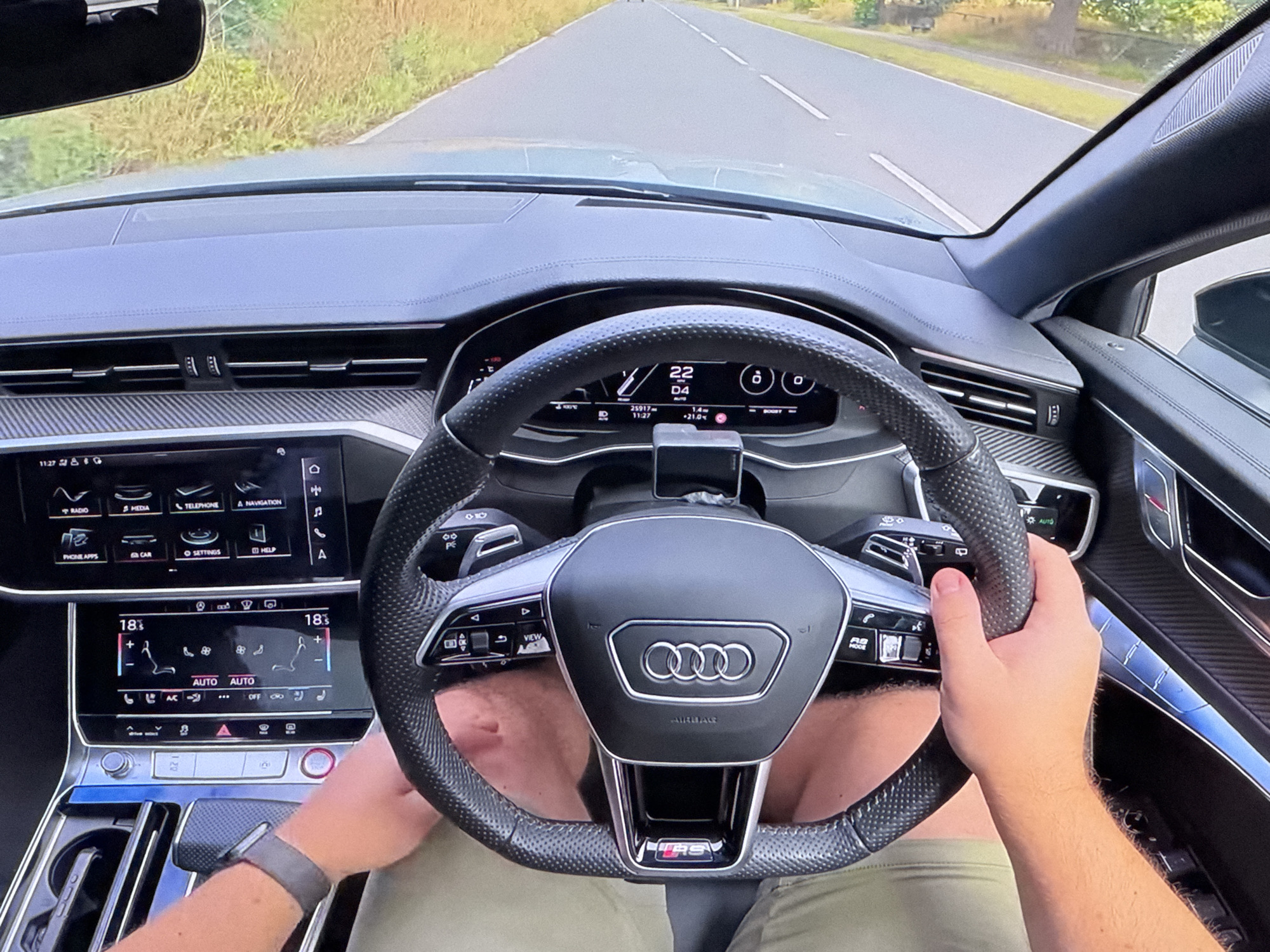 The image shows a first-person perspective from inside a car, specifically an Audi, as it drives down a road. The driver's hands are on the steering wheel, and the dashboard displays are visible, showing navigation and climate control information. The road is paved and flanked by green vegetation. The interior of the car features modern digital displays and carbon fiber accents. There is no visible indication of the car being in Bezden, Bulgaria.