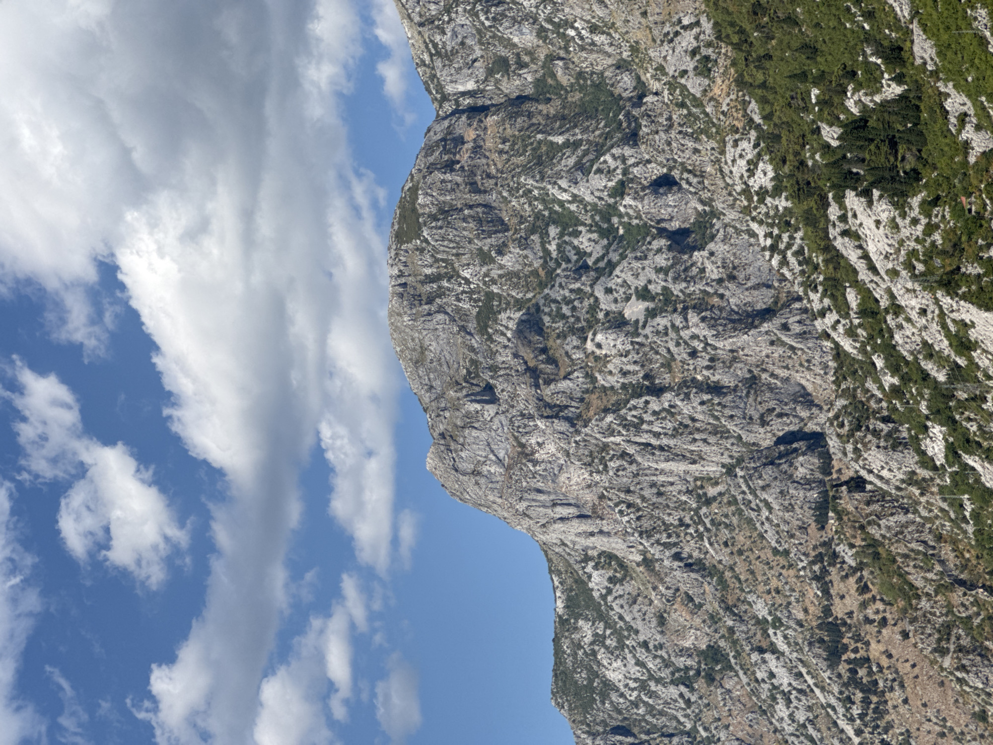 This is a vertical, medium shot photograph of a rocky mountain range under a partly cloudy blue sky. The mountain slopes are rugged and textured, with visible rock formations and sparse vegetation. The lower portion of the image shows more dense greenery, including evergreen trees. The sky above is a vibrant blue with scattered white clouds. The scene depicts a natural, mountainous environment, likely in the vicinity of Škaljari, Montenegro, as indicated by the typical terrain and vegetation of the Balkan region. There are no people or discernible man-made structures clearly visible in this shot, focusing solely on the geological and atmospheric elements of the landscape.