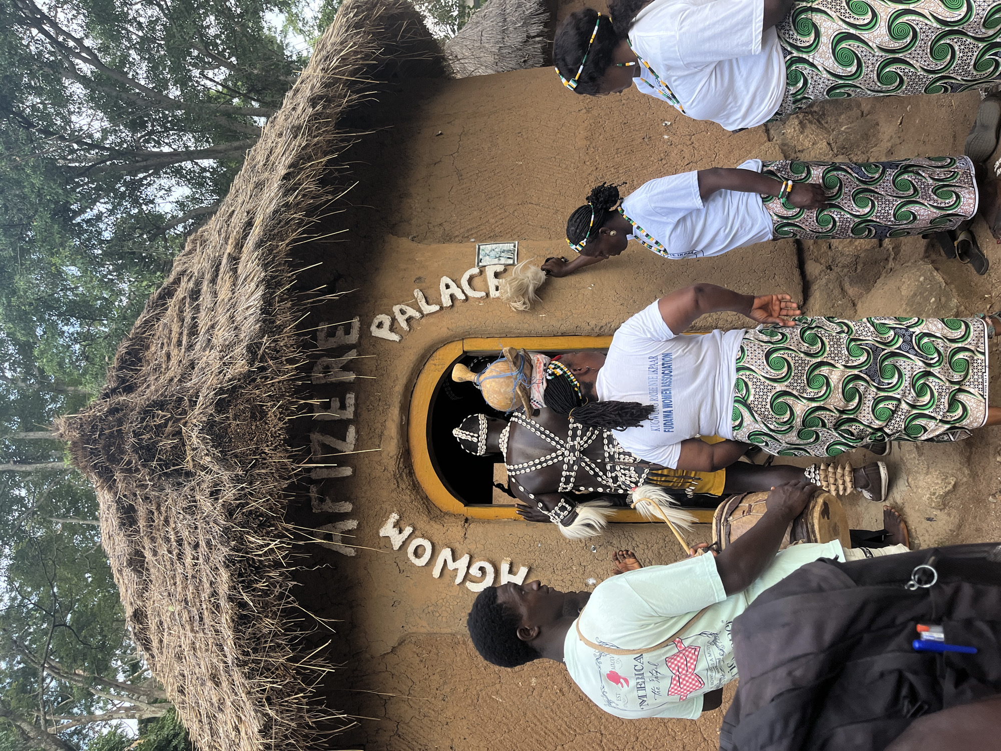 In Jos, Nigeria, a group of people are gathered outside a traditional mud structure with a thatched roof. The structure has text written on its exterior in white: "AFIZERE PALACE" and "HGWOM". Three women, dressed in white t-shirts and patterned skirts featuring green swirls on a white background, are interacting with the building. One woman is touching the wall near a small framed picture. Another woman is leaning in, perhaps speaking or observing. The third woman, in the foreground, is also in a white t-shirt and a patterned skirt.
Inside the doorway of the palace, a person adorned in a traditional outfit with beadwork and cowrie shells is visible. This individual appears to be the focus of attention. A man in a light green t-shirt and shorts is seated and playing a drum, while another person, also in a light green t-shirt with text and graphics, is standing nearby. The overall scene suggests a cultural event or ceremony taking place. The foliage in the background indicates an outdoor setting, likely during daylight hours. The people's attire and the architectural style point to a West African cultural context. Visible text on a t-shirt reads "KUGWUNG RICHE NYE IKPAAR FUDAWA WOMEN ASSOCIATION". Another t-shirt in the foreground has text and graphics that include "MENCA", "The Vetiful", and "SA MUSTA".