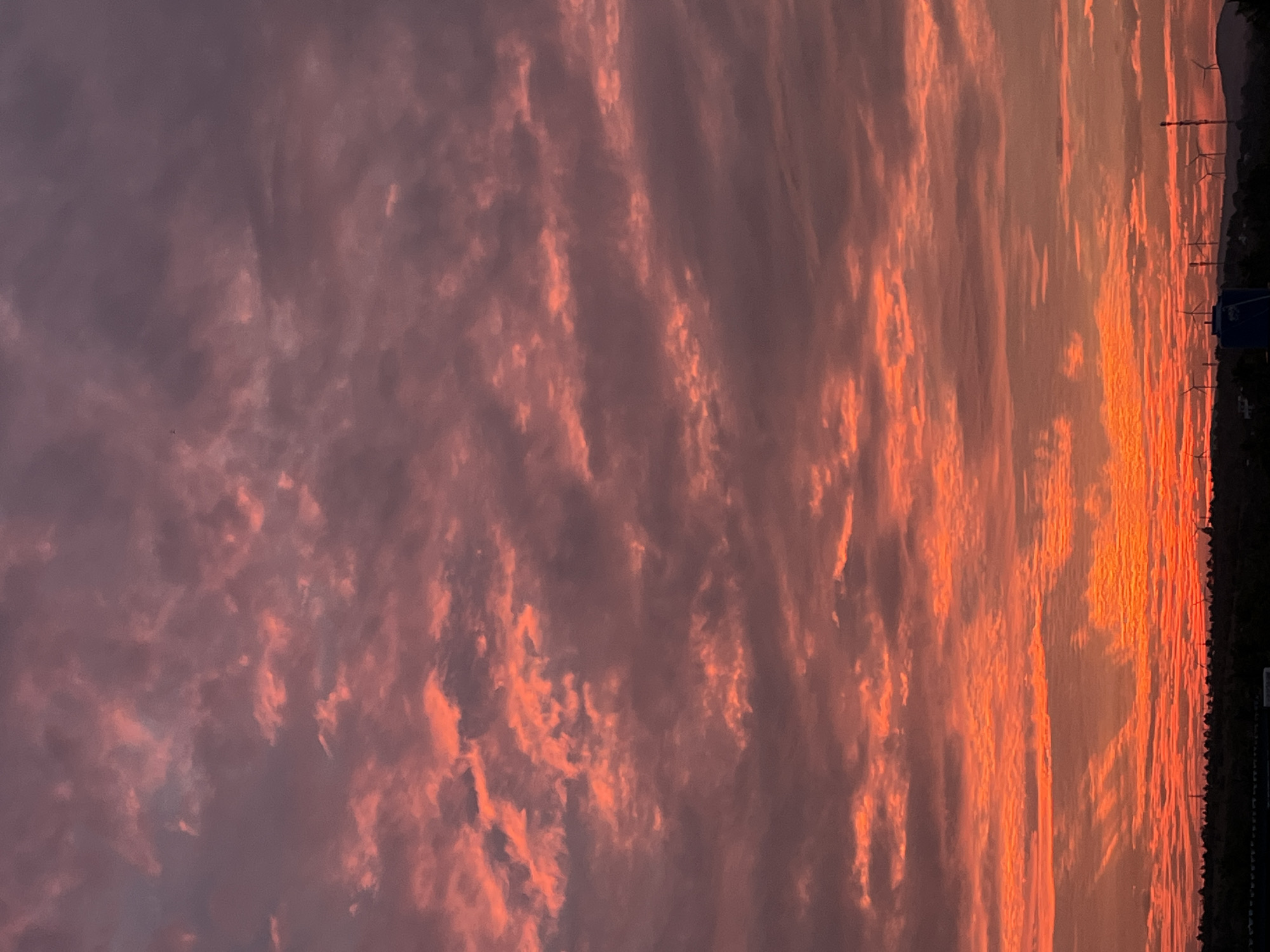 The image depicts a dramatic sunset over Mũi Né, Vietnam. The sky is filled with a vibrant spectacle of clouds, painted in fiery oranges and soft pinks, transitioning to deep purples and grays towards the top of the frame. The clouds are textured and scattered, creating a sense of depth and movement.
Below the sky, a dark silhouette of the landscape stretches across the horizon. This includes the outlines of rolling hills and a dense line of trees. Dotted along the hills are several wind turbines, their tall structures clearly visible against the twilight sky, indicating an area with renewable energy infrastructure. A large blue water tank is also present in the foreground, to the left of center. The overall scene is a serene and powerful depiction of nature's beauty at dusk in the coastal city of Mũi Né.