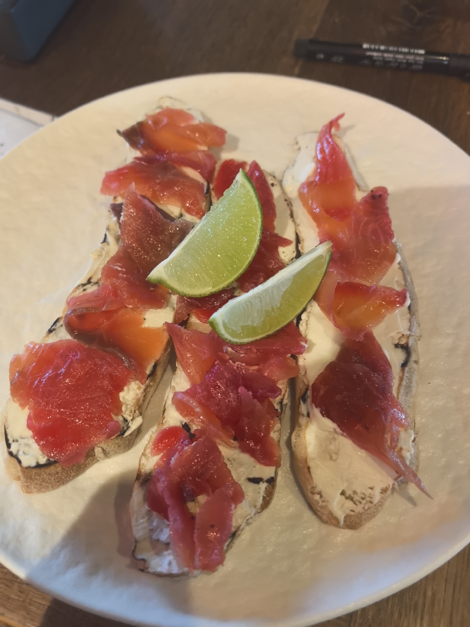 Three open-faced sandwiches are arranged on a white plate. Each sandwich appears to consist of a slice of toasted bread topped with cream cheese and sliced salmon. Two lime wedges are placed on top of the sandwiches. The plate is set on a wooden surface. In the background, there is a glimpse of a document and a black pen. It is impossible to verify from the image whether the location is Varna, Bulgaria.