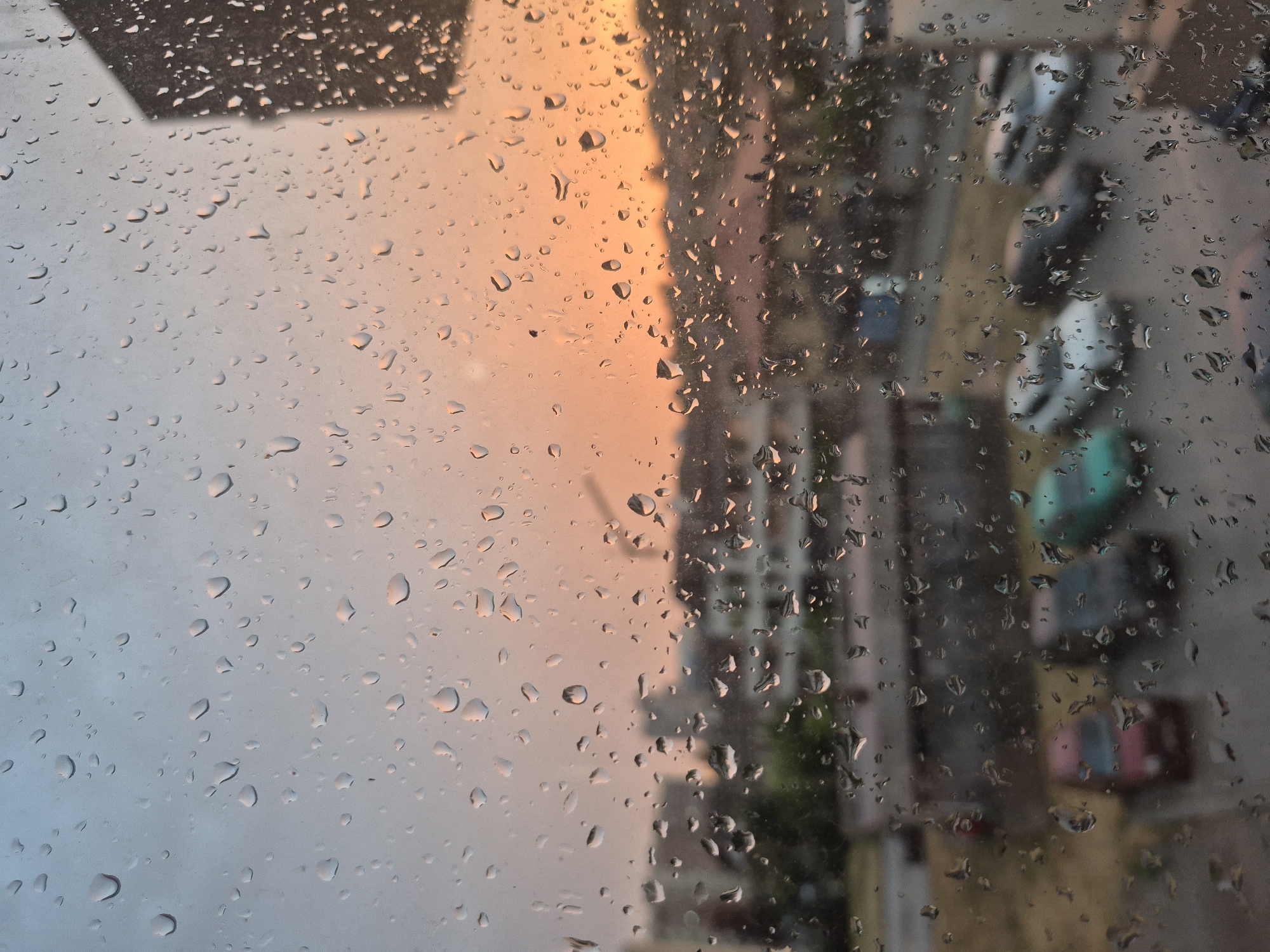 The image captures a view through a rain-streaked window, likely in the city of Popovo, Bulgaria. The foreground is dominated by numerous water droplets of varying sizes clinging to the glass, distorting the scene beyond. A soft, diffused light suggests either dawn or dusk, with the sky transitioning from a pale blue at the top to a warm, peachy orange towards the horizon. In the mid-ground, blurred buildings and structures form the urban landscape. A few cars are visible in a parking area, appearing as indistinct shapes due to the rain and distance. A dark, angular object, possibly a part of a building, juts into the frame from the right side. The overall impression is one of a quiet, possibly overcast day in an urban setting, with the rain adding a layer of texture and mood to the scene.