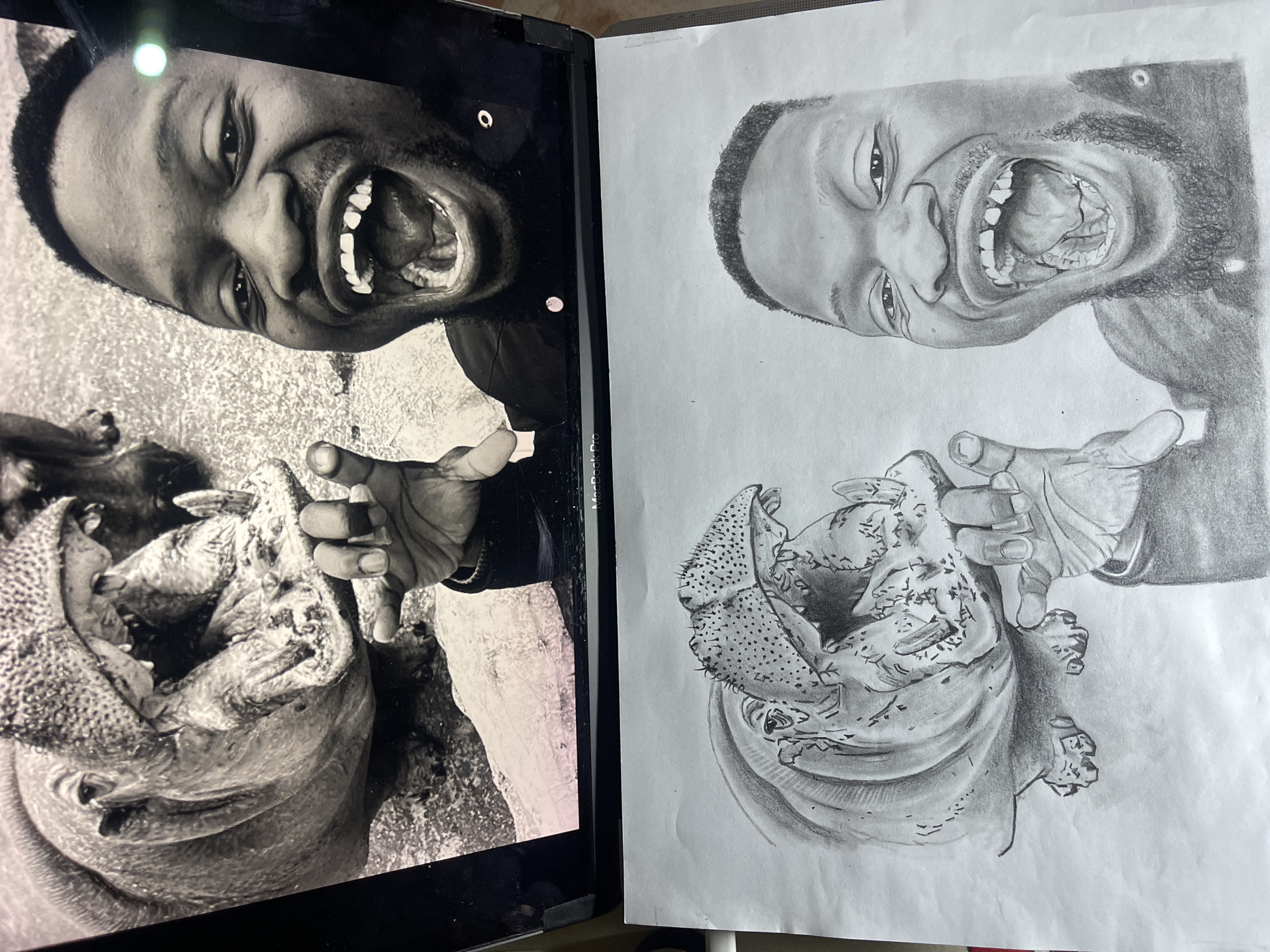 This image displays a MacBook Pro laptop screen positioned above a pencil drawing on white paper. The laptop screen exhibits a black and white photograph featuring two main subjects: an adult male of African ethnicity and a hippopotamus. The man, situated on the right side of the photograph, is captured from the chest up with a wide-open mouth, revealing his tongue, palate, and the absence of his upper central incisors. He has short, dark hair and a short beard, dressed in a dark, long-sleeved garment. His right hand is raised in the foreground, with fingers splayed. The hippopotamus, located on the left, is shown with its mouth agape, exposing prominent tusks and its inner mouth. The man's raised hand is spatially arranged to appear to interact with the hippopotamus's open mouth and tusk, creating a forced perspective effect. The photographic background consists of a textured, possibly outdoor, ground surface.
Directly beneath the laptop, a black and white pencil sketch on white paper meticulously replicates the exact composition and subjects from the photograph. The drawing precisely renders the man's distinctive features, his wide expression, the missing teeth, his raised hand, and the hippopotamus with its open mouth and tusks. The paper itself shows minor undulations. The overall scene suggests an indoor setting, likely a desk or table.
The specific location within the city of Jos, Nigeria, cannot be determined from the visual evidence in the image.
