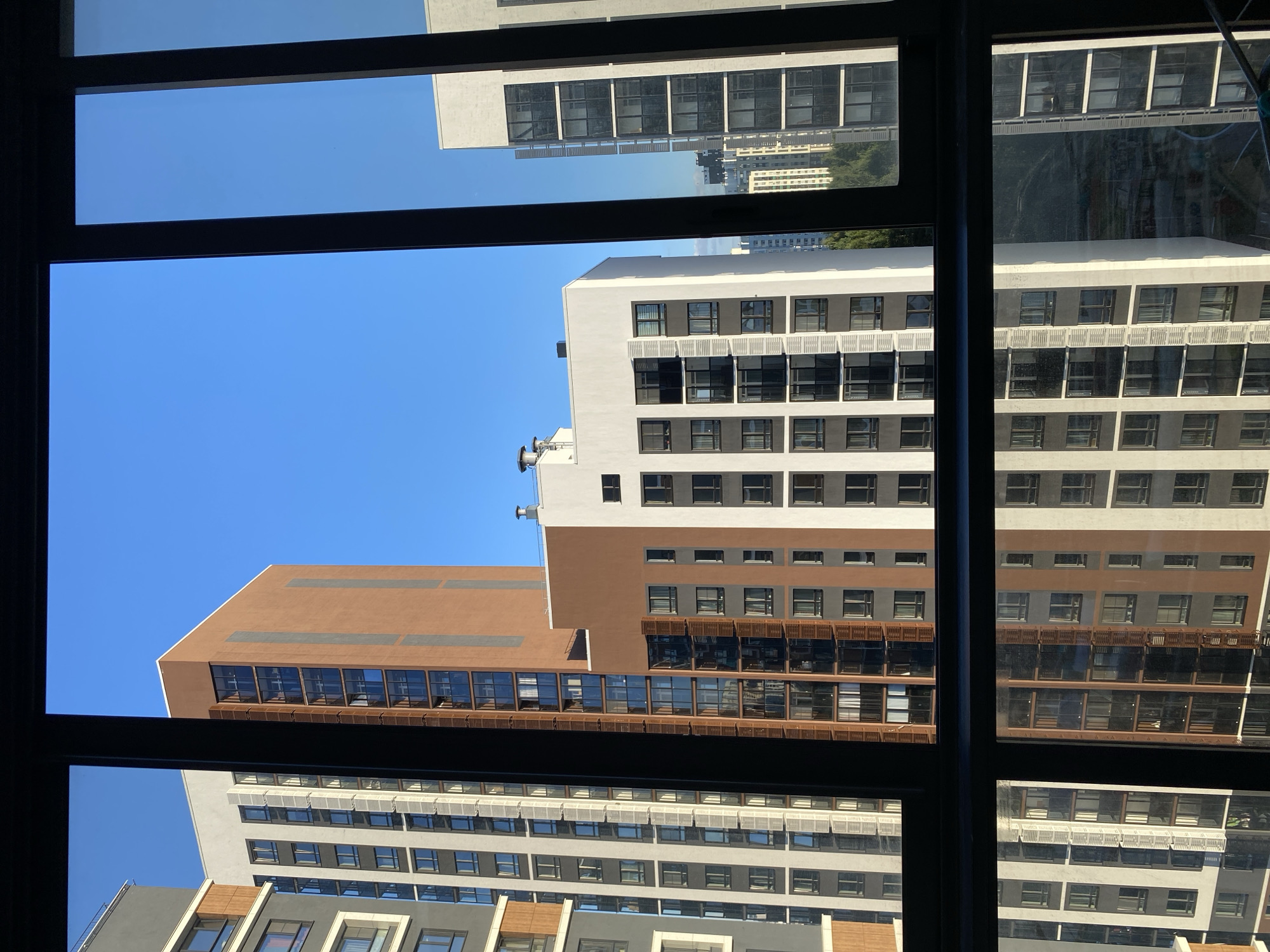 A view from a window shows a clear blue sky above several modern apartment buildings. The buildings are of varying heights and colors, with a mix of white, grey, and brown facades. Some buildings feature balconies and numerous windows. In the foreground, a reflection of these buildings is visible on the glass, creating a mirror image. The scene appears to be in a city, likely Yekaterinburg, Russia, given the architectural style of the buildings. There are no people or objects of note in the foreground, and no discernible actions or interactions are taking place. The environment is urban, characterized by the presence of residential high-rises under a bright, cloudless sky.