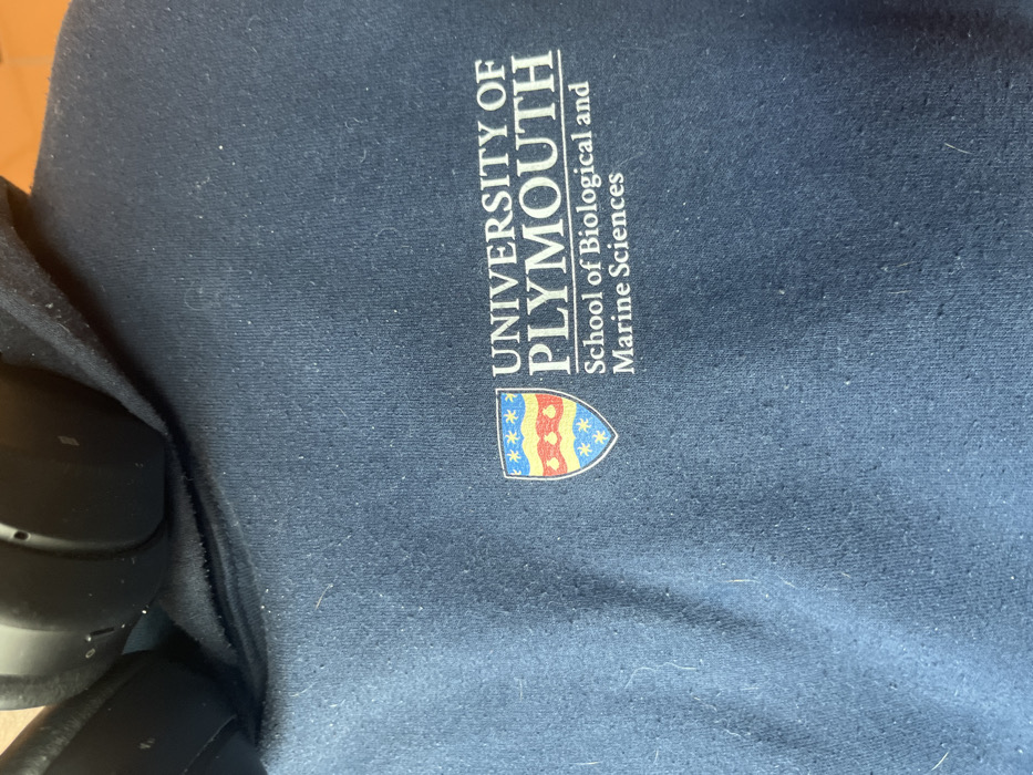 An overhead shot captured at an angle displays a dark blue fabric surface, likely a sweatshirt or hoodie, adorned with the branding of the University of Plymouth. The fabric shows minor pilling and lint, indicating some wear. Text printed in light beige or white is oriented vertically along the right side of the frame, reading "UNIVERSITY OF PLYMOUTH" in a larger, bold font, and beneath it, "School of Biological and Marine Sciences" in a smaller font. To the left of the text, a colorful crest or shield logo is visible, featuring red and yellow wavy stripes, a blue section with white stars, and a solid red section. In the bottom left corner, partially obscured, is a black over-ear headphone, suggesting an indoor setting. The image itself does not provide any specific cues for time of day, weather, or the provided location context of Dorno, Italy. No people are visible.