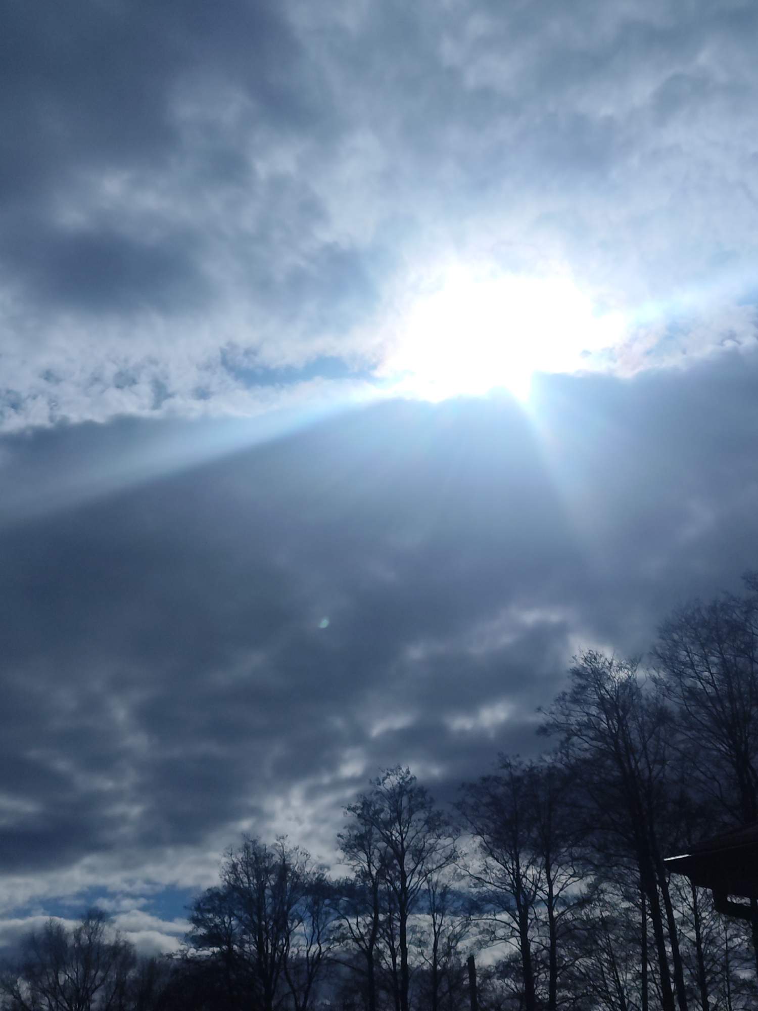 The image shows a cloudy sky with the sun shining through a gap in the clouds. A visible sunbeam is emanating from the sun. In the foreground, silhouettes of trees are visible. The location is unknown.