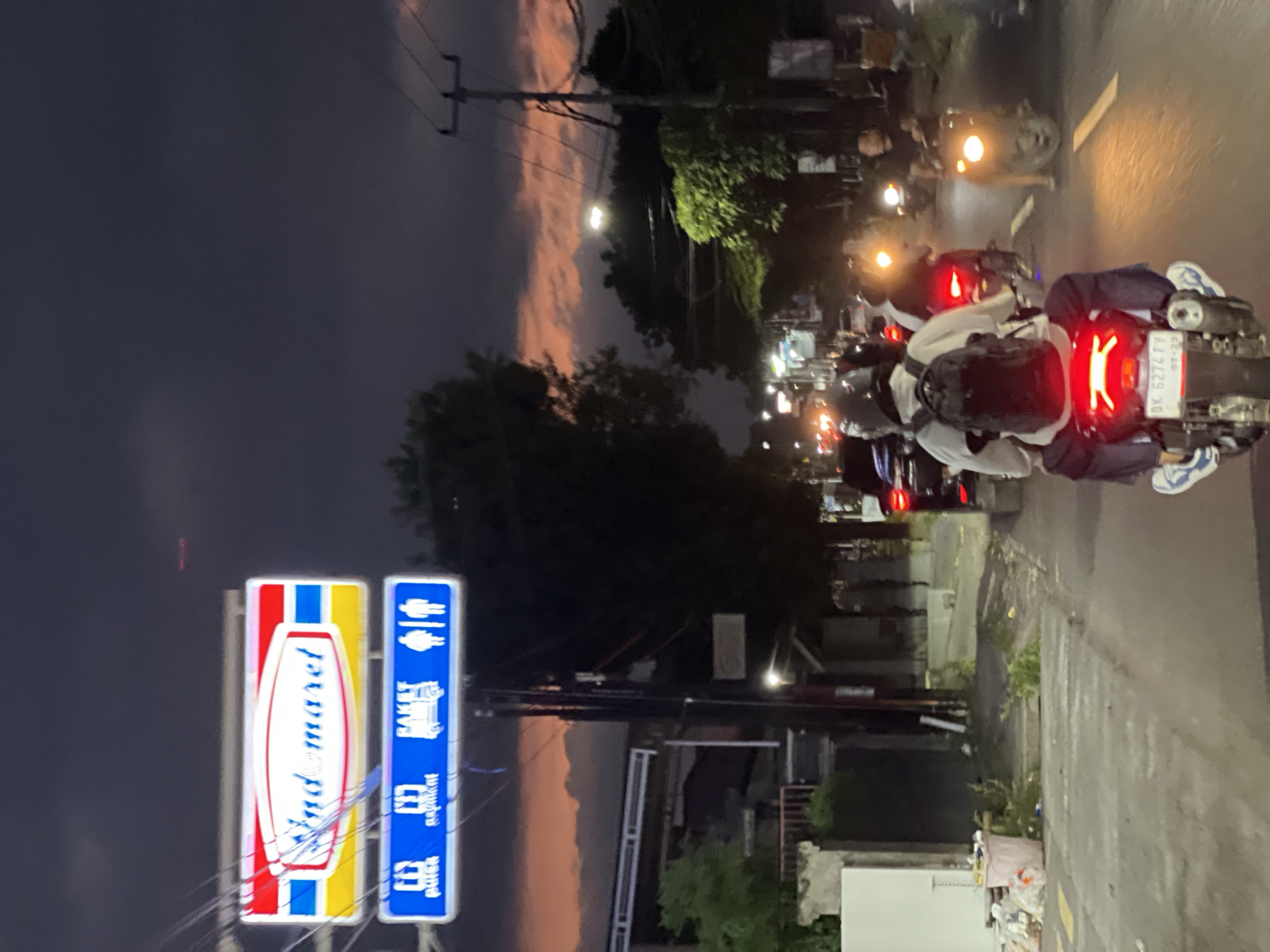 This is a vertical, medium-shot image taken at dusk in Sawan, Indonesia. The scene depicts a street with several motorcycles lined up, some with riders. The motorcycles have their brake lights illuminated, casting a red glow. The street appears wet, suggesting recent rain or high humidity.
In the foreground on the left, there is a brightly lit sign for "Indomaret," a convenience store chain, and another blue sign with pictograms and text, one of which appears to be "Bakers." The sky above is a deep twilight blue, with hints of soft pink and orange clouds, indicating sunset or sunrise. A utility pole with wires stretches across the frame.
In the background, more motorcycles and a few people are visible. The overall atmosphere is one of a street scene at the end of the day, possibly a gathering or a pause in travel. The lighting is dim, with the primary sources being the signs and the motorcycle lights. The wet street reflects the lights, adding to the moody ambiance. The presence of the convenience store sign provides a strong locational cue.