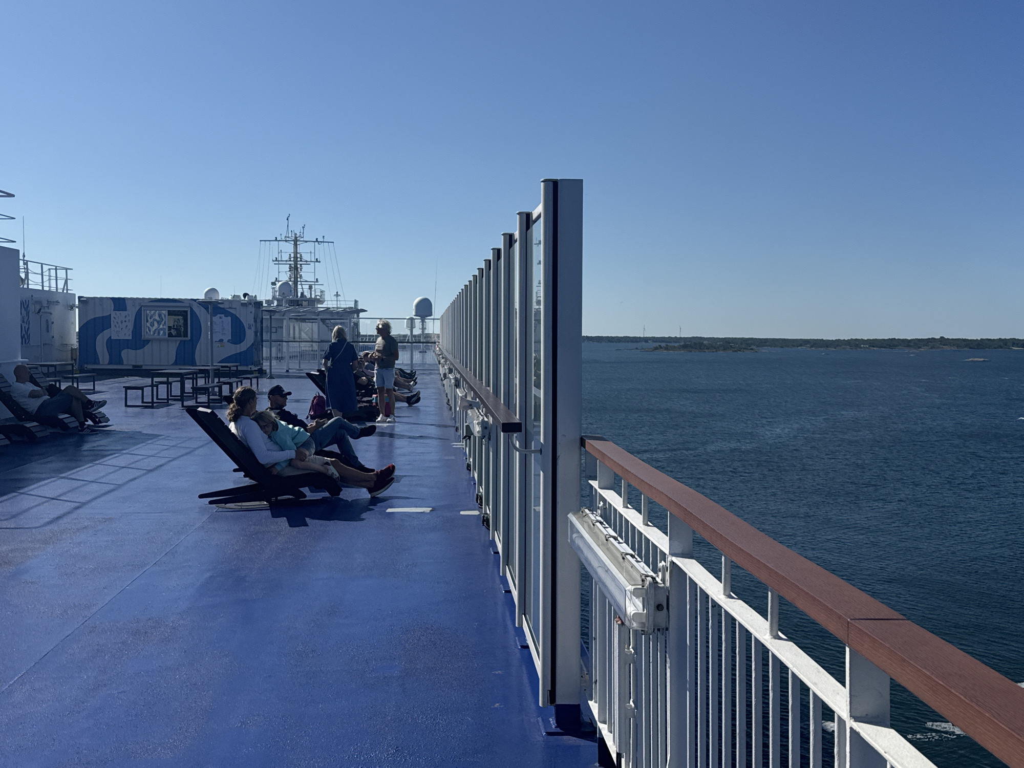The photograph captures an exterior deck of a ferryboat sailing on a clear, sunny day. The deck is painted a vibrant blue, with white railings and a wooden handrail running along the side. Several people are seated on lounge chairs, facing towards the water, and engaged in various activities like reading or relaxing.
In the background, a large white building structure with blue patterns, possibly a shipping container, is visible on the deck. Further beyond that, the superstructure of the ferry, including antennas and navigational equipment, is apparent. The ferry is navigating through a body of calm, deep blue water, with a distant shoreline dotted with trees and wind turbines, suggesting a coastal landscape. The sky is a clear, cloudless blue.
The image has been identified as being taken in Karlskrona, Sweden, a city known for its naval history and archipelago. The ferry is likely traveling between islands or to the mainland, showcasing a typical scene of maritime transport in the region.