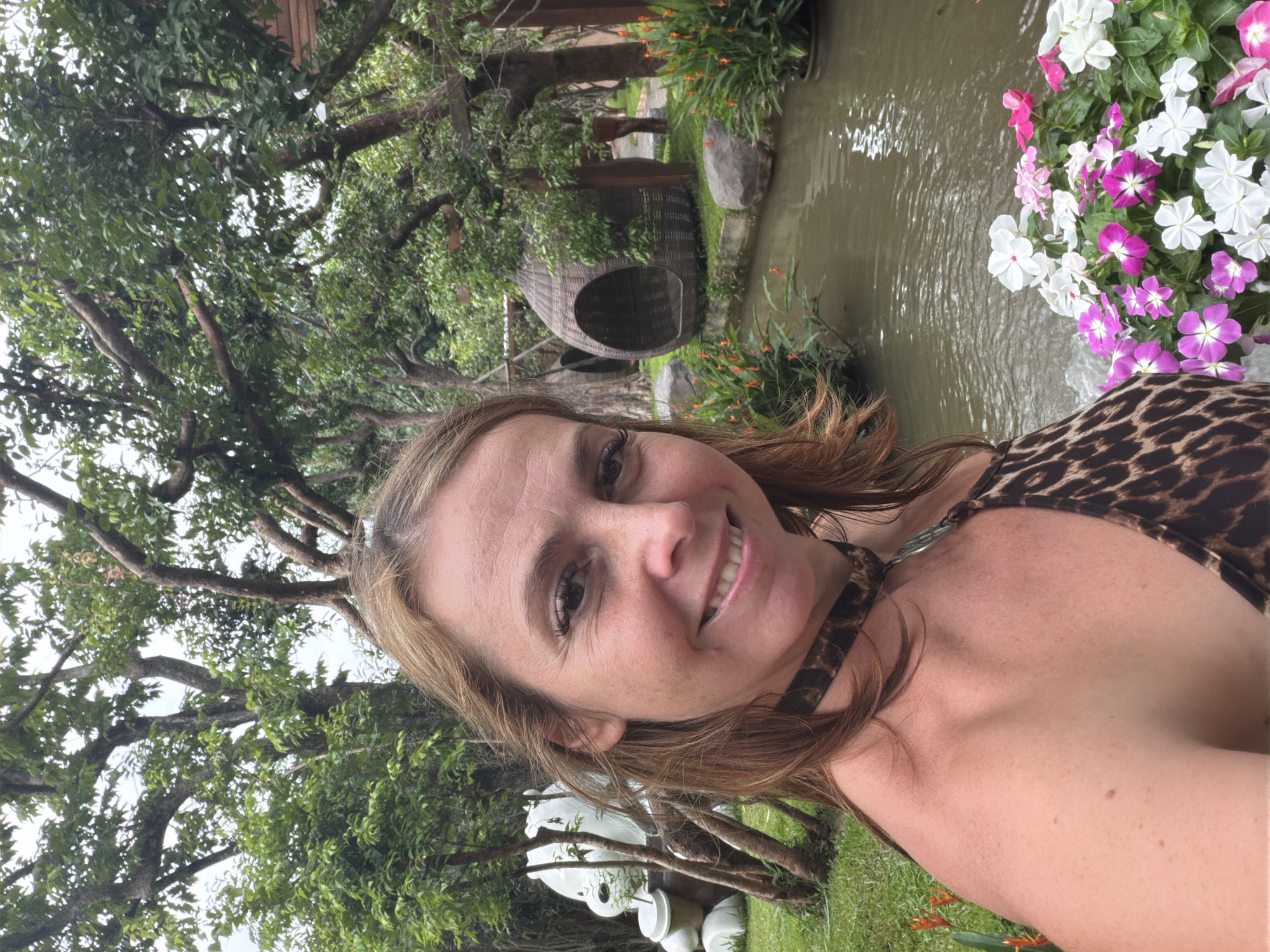 A woman with shoulder-length brown hair smiles at the camera in a selfie. She is wearing a leopard-print bikini top and a black choker necklace. Behind her, a small pond is surrounded by lush greenery and trees. A woven pod-shaped chair hangs from a tree branch over the pond. In the background, a large white bear sculpture with a cup is visible. The scene is outdoors, with a cloudy sky overhead, suggesting it might be in Tambon Mae Raem, Thailand, a region known for its natural beauty and tourist attractions.
