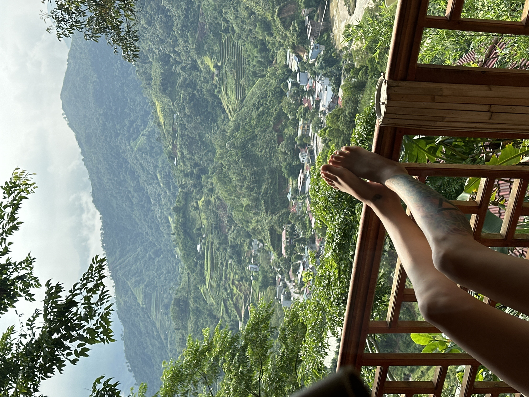 The image captures a serene, expansive view of a mountainous landscape and a village in Thông Nguyên Commune, Vietnam, as seen from a balcony or terrace. In the immediate foreground, a person's bare legs and feet are propped up on a rustic wooden railing, suggesting relaxation. A distinct tattoo is visible on the calf of the right leg.
Beyond the railing, a deep valley unfolds, densely covered in vibrant green foliage and trees. A village is nestled within this valley, featuring numerous houses with various rooftops scattered across the landscape. Further up the mountain slopes, well-defined agricultural terraces, likely rice paddies, are visible, showcasing the area's agricultural practices. The background is dominated by towering, verdant mountains, their peaks partially obscured by a partly cloudy or hazy sky.
The scene is illuminated by bright daylight, indicating it was taken during the day. The overall impression is one of tranquility and natural beauty, with a blend of human settlement and untouched nature. Lush green plants and trees frame the view from the foreground. No visible text is present in the image.