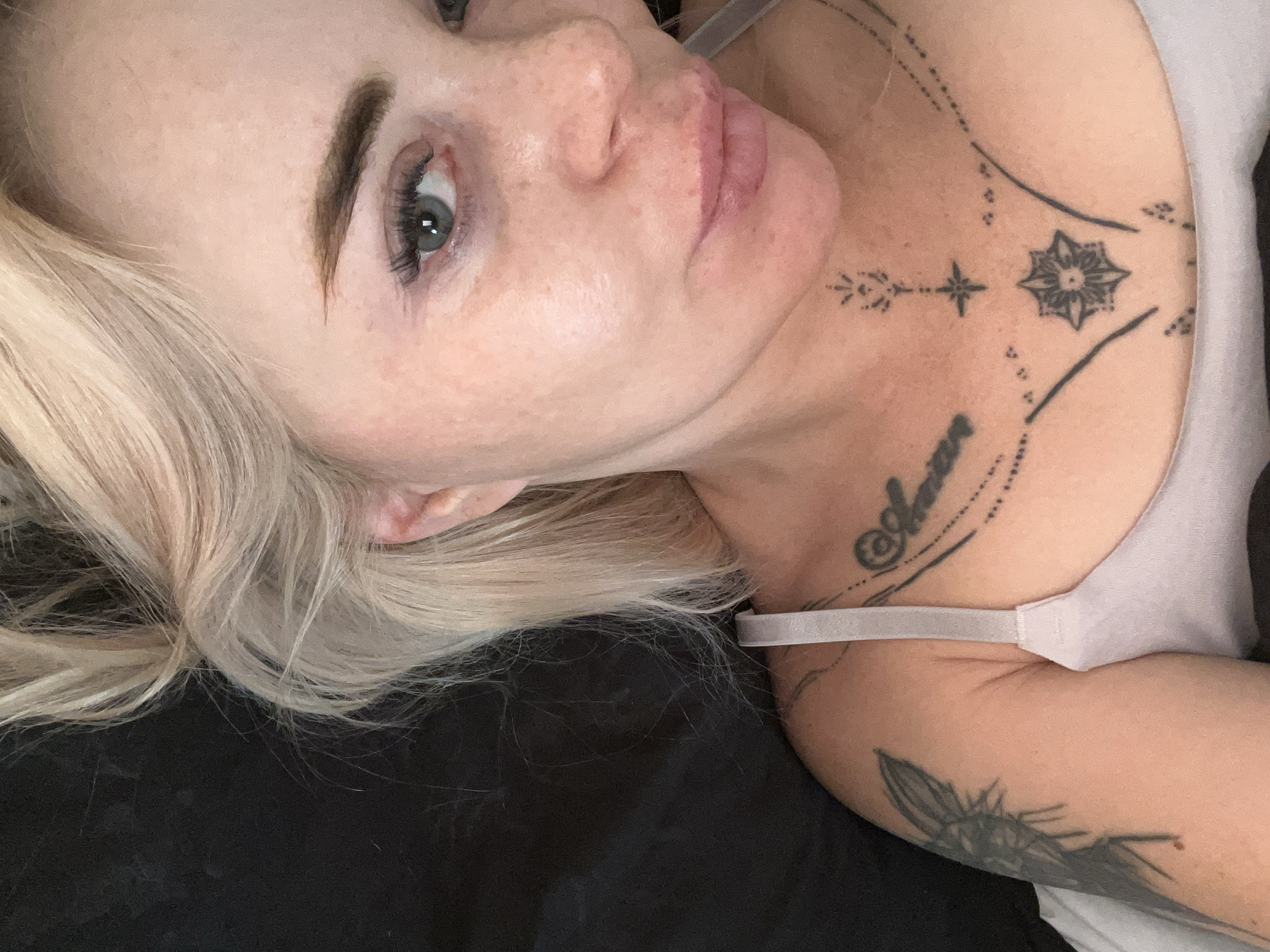 A close-up, head-and-shoulders shot captures a person with fair skin, blonde hair, and light blue eyes. Their face is turned towards the camera, with their gaze directed slightly off-frame. The subject has freckles across their nose and cheeks, and their lips are slightly parted. They are wearing a light-colored bra, with a strap visible across their chest. There are several tattoos visible on the subject, including one on their left arm and intricate designs on their chest and neck. The background is dark and appears to be a bed or similar surface. The lighting is soft, illuminating the subject's face. No specific location in a city is discernible from the image.