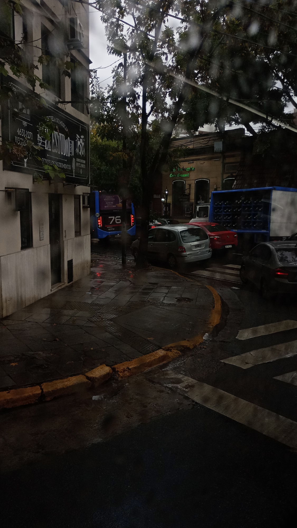 The image depicts a street scene in Buenos Aires, Argentina, on a rainy day. The location features a building on the left with a sign indicating "Escuela de Música" (Music School), with the phone numbers 4631-1271 and 113316. A bus with the number 76 and an advertisement for Shen Yun is visible, along with a silver hatchback, a red sedan, and a box truck. A building across the street has a sign that says "Bohemia Colly Concert". The street is wet and there are crosswalk markings and yellow curbs visible in the foreground.