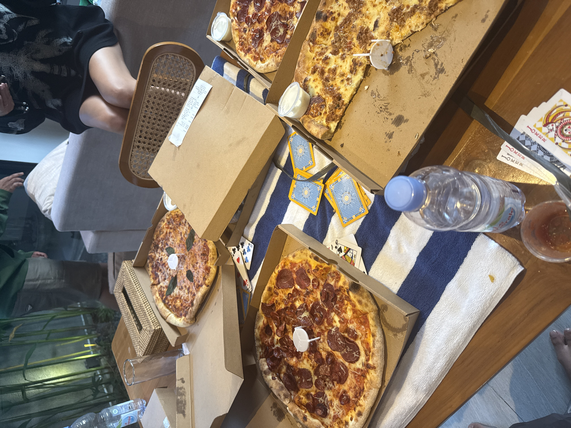 In Mengwi, Indonesia, a casual gathering is taking place, possibly at a home or resort, with several pizzas, playing cards, and drinks laid out on a table and on a striped towel. Two large pizzas in open cardboard boxes are prominent. One is topped with pepperoni and basil, while the other appears to be a meat lover's pizza. Two smaller pizzas are also visible, one with pepperoni and another with cheese and basil. Small containers of dipping sauce are next to some of the pizzas. Several decks of playing cards are scattered among the food, suggesting a game is in progress or has recently concluded. A plastic water bottle and a glass are also on the table. In the background, a person in a green sweatshirt and shorts stands near lush green plants, and another person is seated on a sofa, wearing a black dress. The overall atmosphere suggests a relaxed, social occasion.