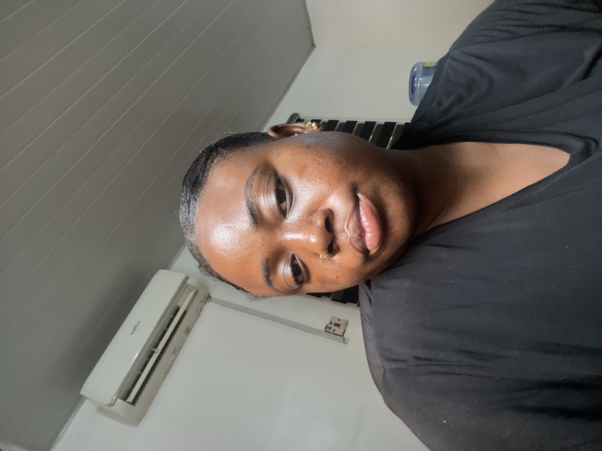 A close-up, low-angle shot of a Black woman with dark hair pulled back, wearing a black top. She has a gold ring in her left nostril and gold earrings. The background shows a room with a horizontally-paneled white ceiling, a white air conditioning unit mounted on a white wall, and a power outlet. A blue water bottle is partially visible near the top of the frame. The location appears to be indoors, possibly in Abuja, Nigeria.