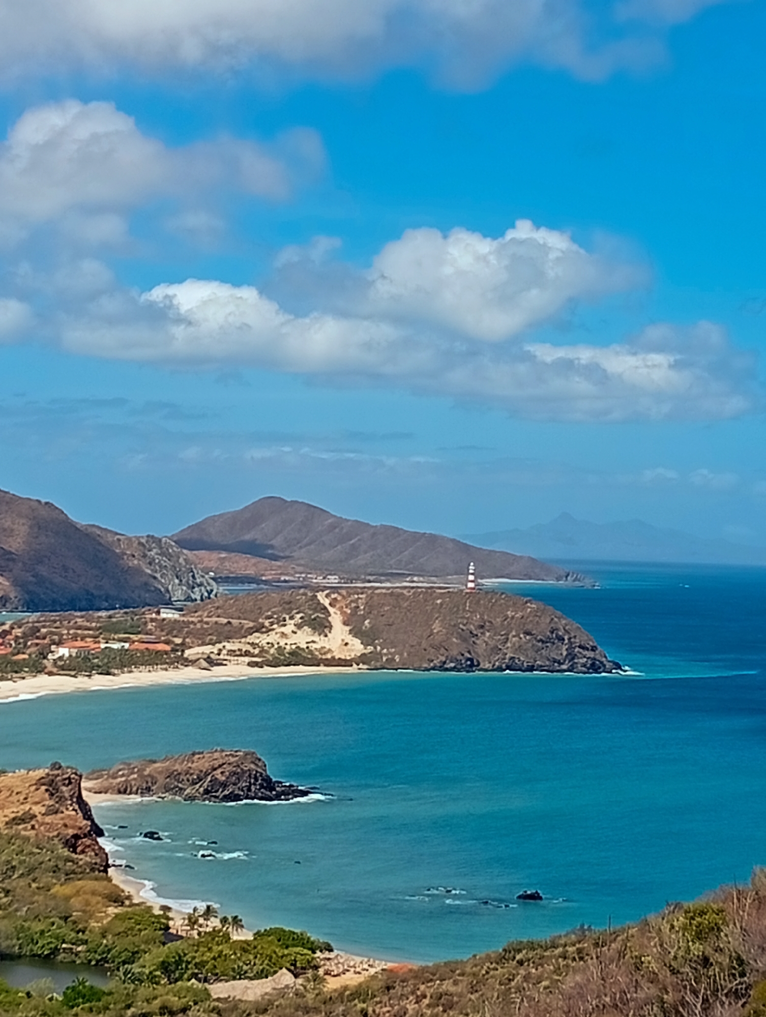 The image displays a coastal panorama near Juan Griego, Venezuela, under a bright blue sky dotted with scattered white cumulus clouds. The foreground consists of dry, rocky hillsides covered with sparse, light-brown and green vegetation, including cacti. In the lower left, a tranquil body of dark-green water, likely an inlet or lagoon, is visible, bordered by denser green foliage and palm trees. The midground features several bays with clear turquoise-blue water and light sandy beaches. Along one beach, multiple buildings with red-tiled roofs are visible. A prominent brown rocky headland extends into the sea, topped by a red and white striped lighthouse. Beyond this, the deeper blue ocean extends to the horizon, where hazy, distant mountains are faintly discernible. Larger, arid brown mountains form the backdrop to the coastal plain. Gentle waves are observed breaking against the shorelines and rocky outcrops. No individuals or active watercraft are present in the scene.