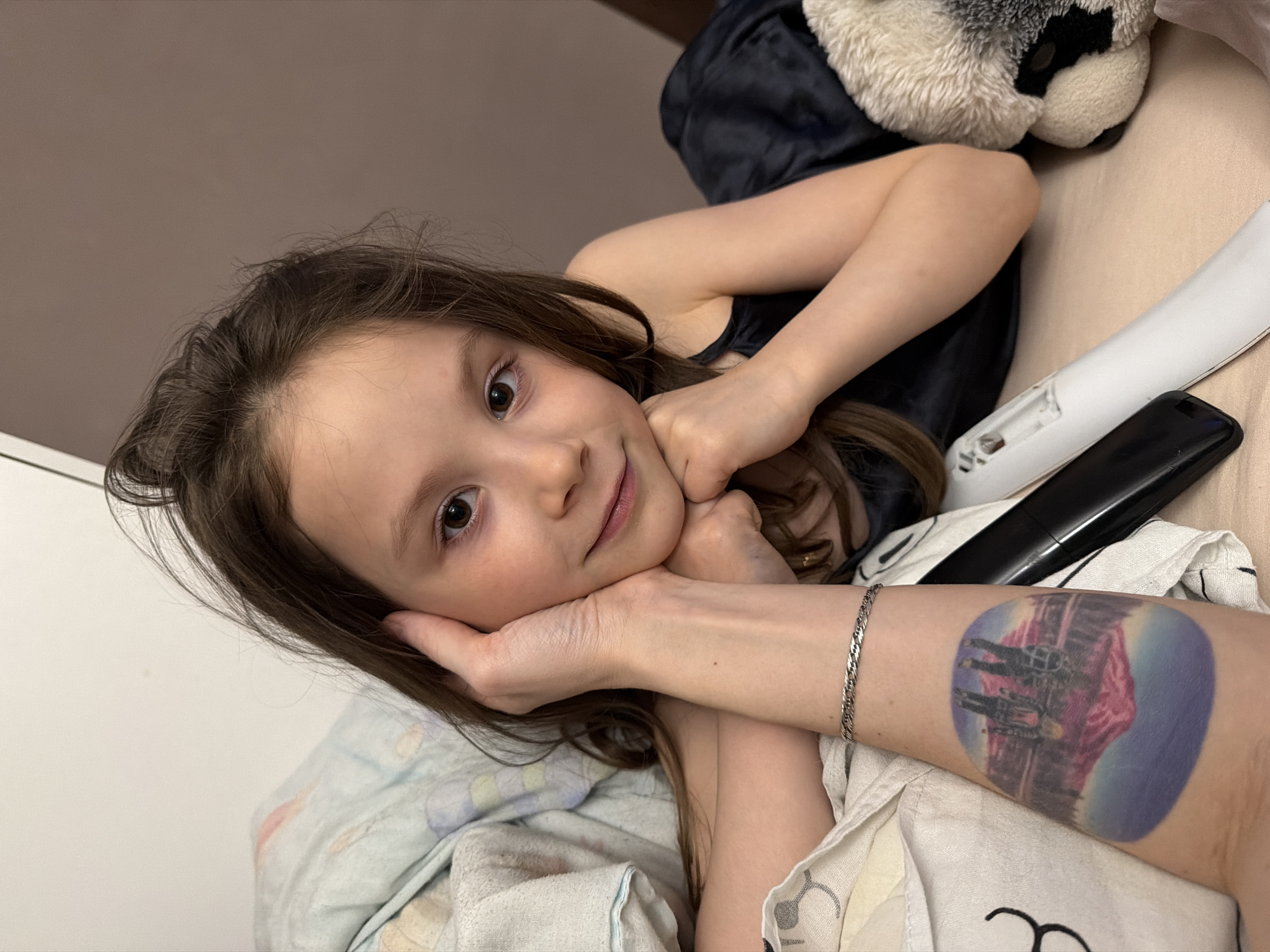 A young girl, estimated to be between 5 and 8 years old, is depicted in a close-up, slightly tilted portrait, resting her head on an adult's hand. She has brown hair and brown eyes, looking directly at the camera with a soft, contented smile, and is wearing a dark, sleeveless top.
The adult's arm, presumably female, supports the girl's chin. A silver braided bracelet adorns the adult's wrist, and a striking, colorful tattoo is visible on her forearm. The tattoo depicts a cityscape, possibly with reflections in water, under a gradient sky of purple and blue hues.
The scene takes place indoors, likely a bedroom or living room in Perm, Russia. The background features a beige or light brown wall/surface on the right and a white wall on the left. Various household items are scattered around the girl: a white remote control with its battery compartment exposed (missing cover), a black remote, and a black smartphone are visible on the right. A fluffy white and black stuffed animal can be seen in the upper right corner. The girl is partially covered by light-colored bedding, including a patterned blanket and a white, towel-like fabric. The overall mood of the image is intimate and relaxed, capturing a warm moment between the child and the adult. No visible text is present in the image.