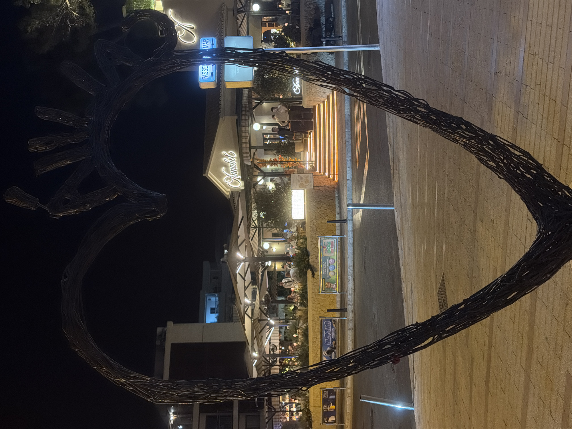 A large, dark, wire-frame heart sculpture is positioned in the foreground, obscuring the lower part of the scene. Behind the sculpture, a well-lit restaurant named "Annabel" is visible. The restaurant has an outdoor seating area with white umbrellas and patrons dining. Stairs leading up to the restaurant entrance are illuminated with warm, orange lights. Two blue street signs are mounted on a pole, one reading "PASSEIG" and the other "MAR". The area appears to be a pedestrian walkway or promenade. In the background, there are multi-story buildings. The time appears to be night, indicated by the dark sky and artificial lighting. The city is Palmanova, Spain.