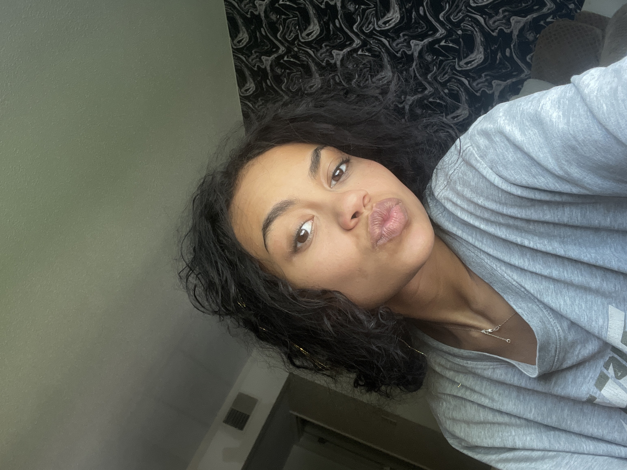 A young woman with dark, curly hair and brown eyes is shown in a close-up, selfie-style shot. She is wearing a grey t-shirt and a delicate silver necklace. Her lips are pursed, giving a playful expression. The background features a wall with a black and white marbled pattern, and to the left, a glimpse of a doorway or closet with vertical blinds is visible. The lighting suggests it is daytime, with light coming from the right. The scene appears to be indoors, likely in a bedroom or living space. No specific location in Boone, United States of America, can be determined from the image alone.