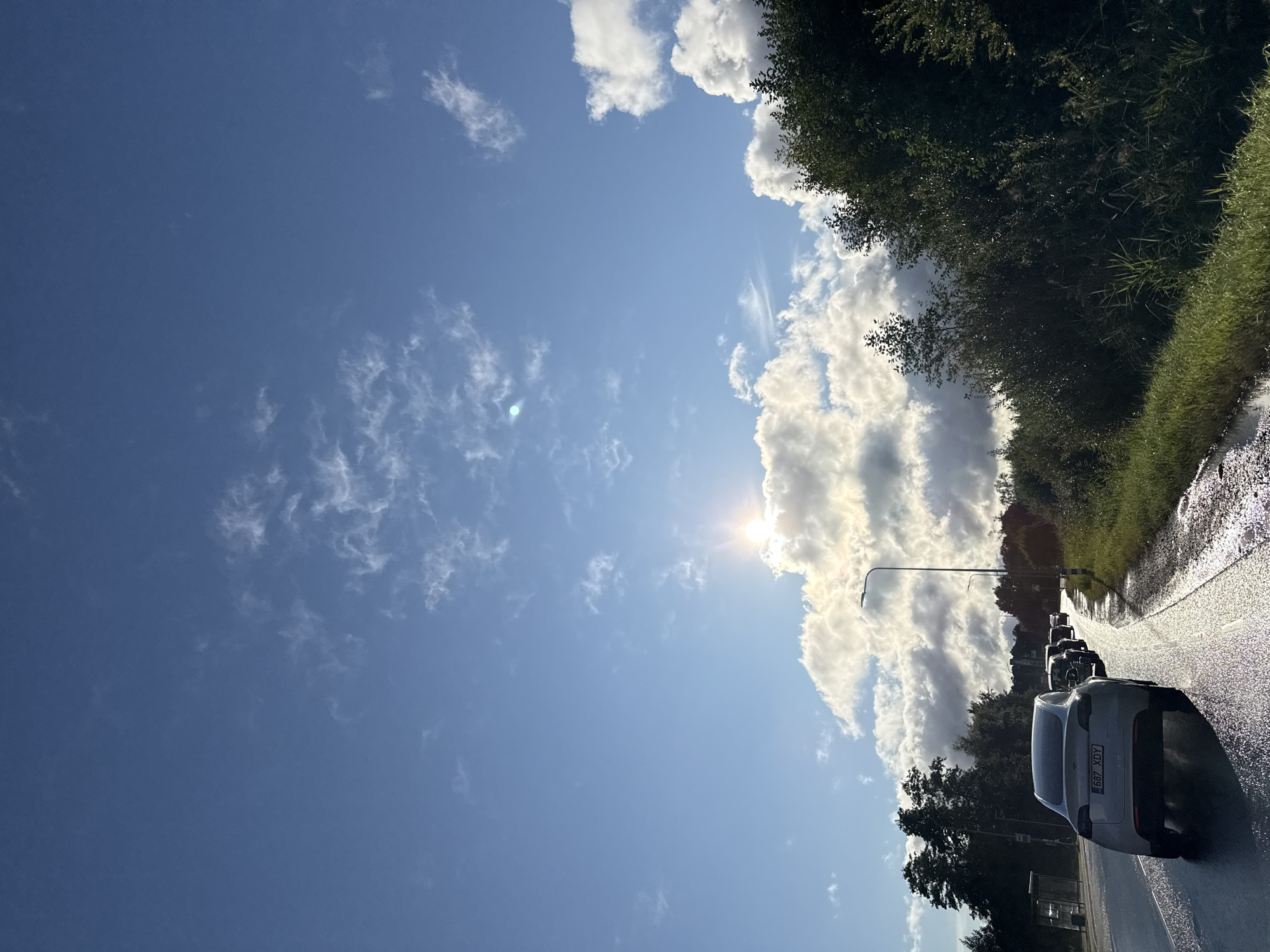 This is a photograph taken from a vehicle traveling on a wet road in Tallinn, Estonia, under a bright, partly cloudy sky. The sun is partially obscured by clouds, casting a glare. The road appears to be a multi-lane highway or main road, with a line of cars in front of the camera. The vehicle in the foreground is a white Tesla, with a license plate visible. To the right of the road, there is a dense line of trees and vegetation. To the left, there is a sidewalk and a bus stop with a shelter. The overall scene suggests a journey through a suburban or semi-rural area on a day with mixed weather conditions.