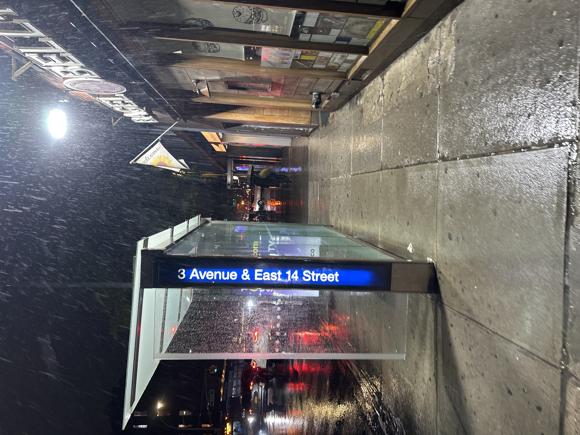 The image depicts a rainy night scene at the intersection of 3rd Avenue and East 14th Street in New York City. A subway entrance sign for the 3rd Avenue and East 14th Street station is prominently displayed, illuminated with blue light. The street and sidewalk are wet from the rain, reflecting the city lights and car headlights. Raindrops are visible, indicating active rainfall. The scene includes glimpses of surrounding buildings, street signs, and vehicular traffic in the background.