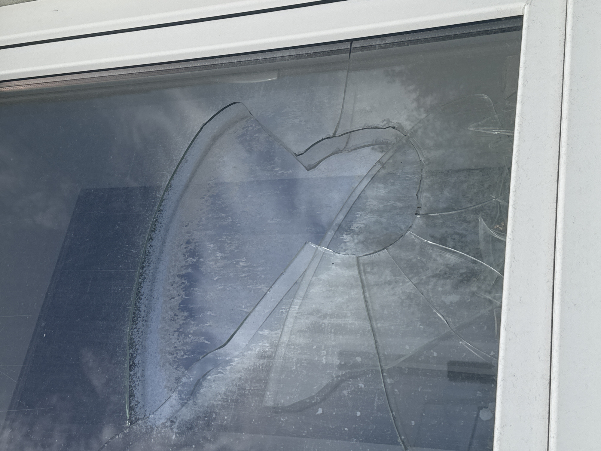 The image shows a close-up of a broken window in Tallinn, Estonia. The glass is cracked and shattered into multiple pieces, with a large section missing from the top right. The window frame is white and appears to be made of uPVC. The reflection in the glass shows a cloudy sky. The scene is outdoors and appears to be daytime. There are no people visible in the image. The damage to the window suggests it may have been caused by an impact or vandalism. The location in Tallinn is not specified, but the overall context is that of urban decay or property damage.