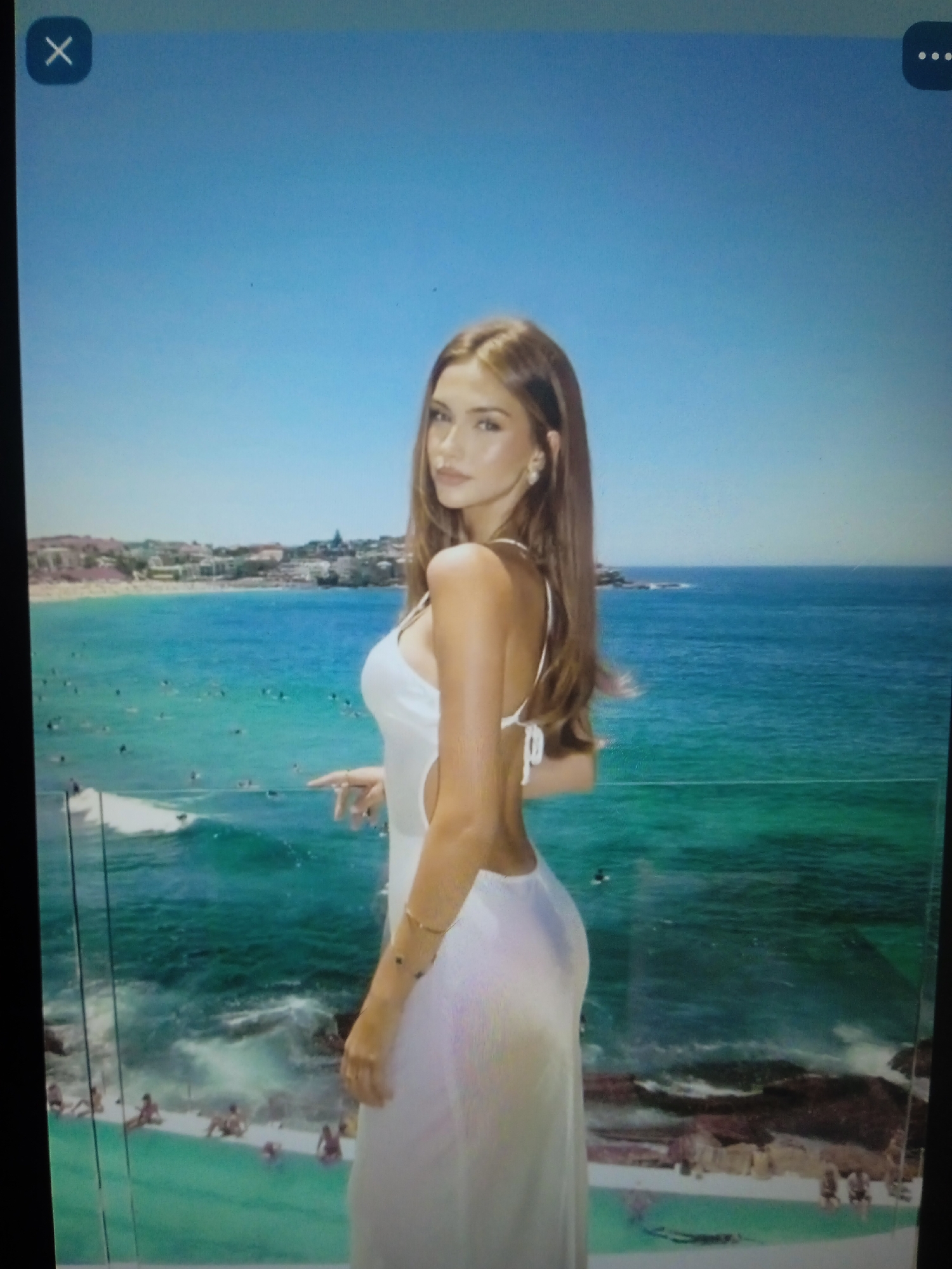 The image shows a young woman in a white dress standing on a balcony overlooking a beach and the ocean. She is facing away from the camera and looking out at the water. The woman has long brown hair and is wearing earrings. The dress is sleeveless and has a low back. The beach is crowded with people, and there are several surfers in the water. The ocean is a vibrant blue color, and the sky is clear and sunny. The buildings in the background suggest an urban coastal setting. There are no elements in the image that specifically indicate the location is Chernivtsi, Ukraine.