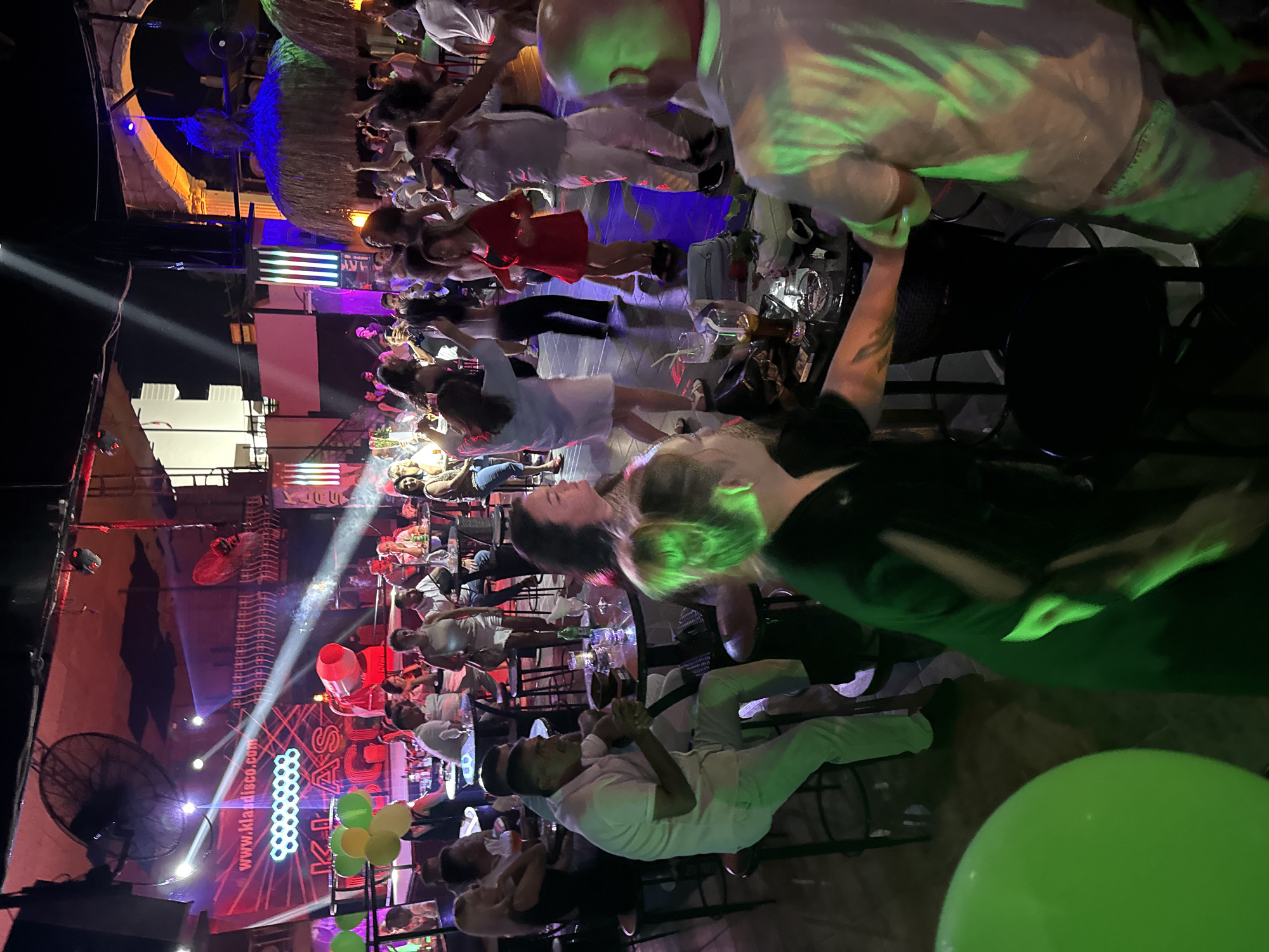 The image depicts an indoor nightlife scene at "Klas Disco" in Alanya, Turkey. The venue appears to be a disco or nightclub with a dance floor and seating areas. Multiple people are present, some dancing and others seated at tables. There are colored lights illuminating the space, and a beam of light shines down from the ceiling. In the background, a large screen displays the website "www.klasdisco.com" along with other graphics. Balloons, some green and yellow, are visible. The atmosphere suggests a lively evening with music and social interaction.