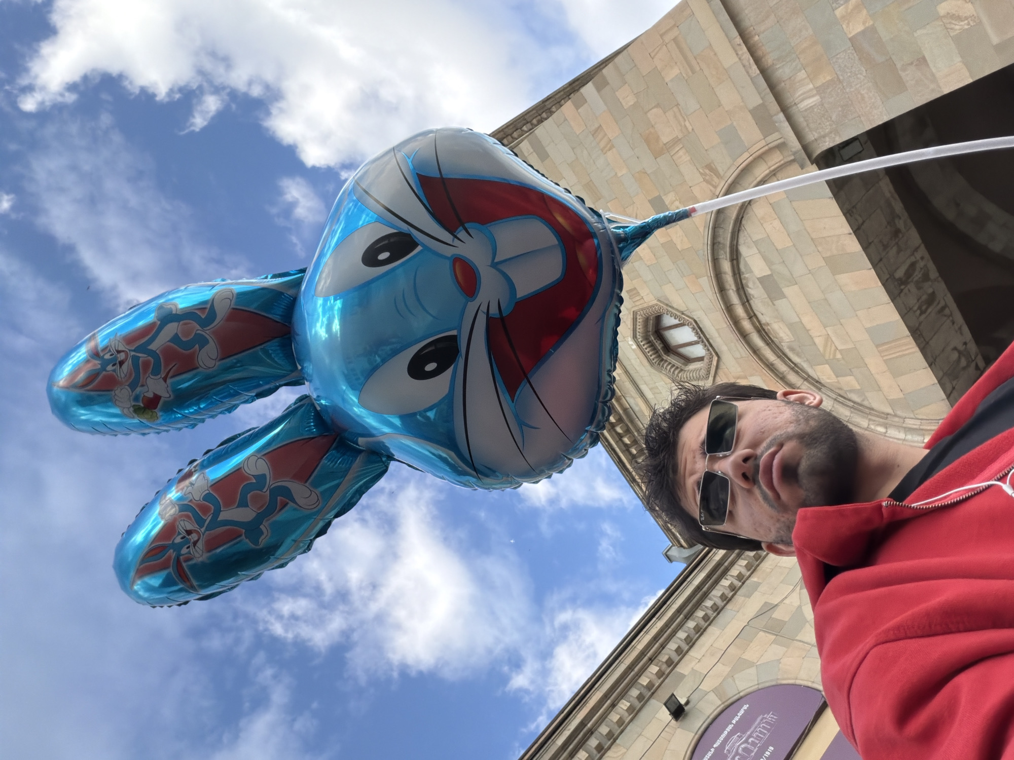 The image shows a man holding a large, inflated balloon shaped like a cartoon character, possibly Doraemon or similar, with rabbit ears featuring Bugs Bunny, against a backdrop of a cloudy sky and a stone building. The man wears sunglasses and a red hoodie. The building is constructed of light-colored stone, with architectural details suggesting a historic or governmental structure.
The architecture of the building suggests the location is in Yerevan, Armenia.