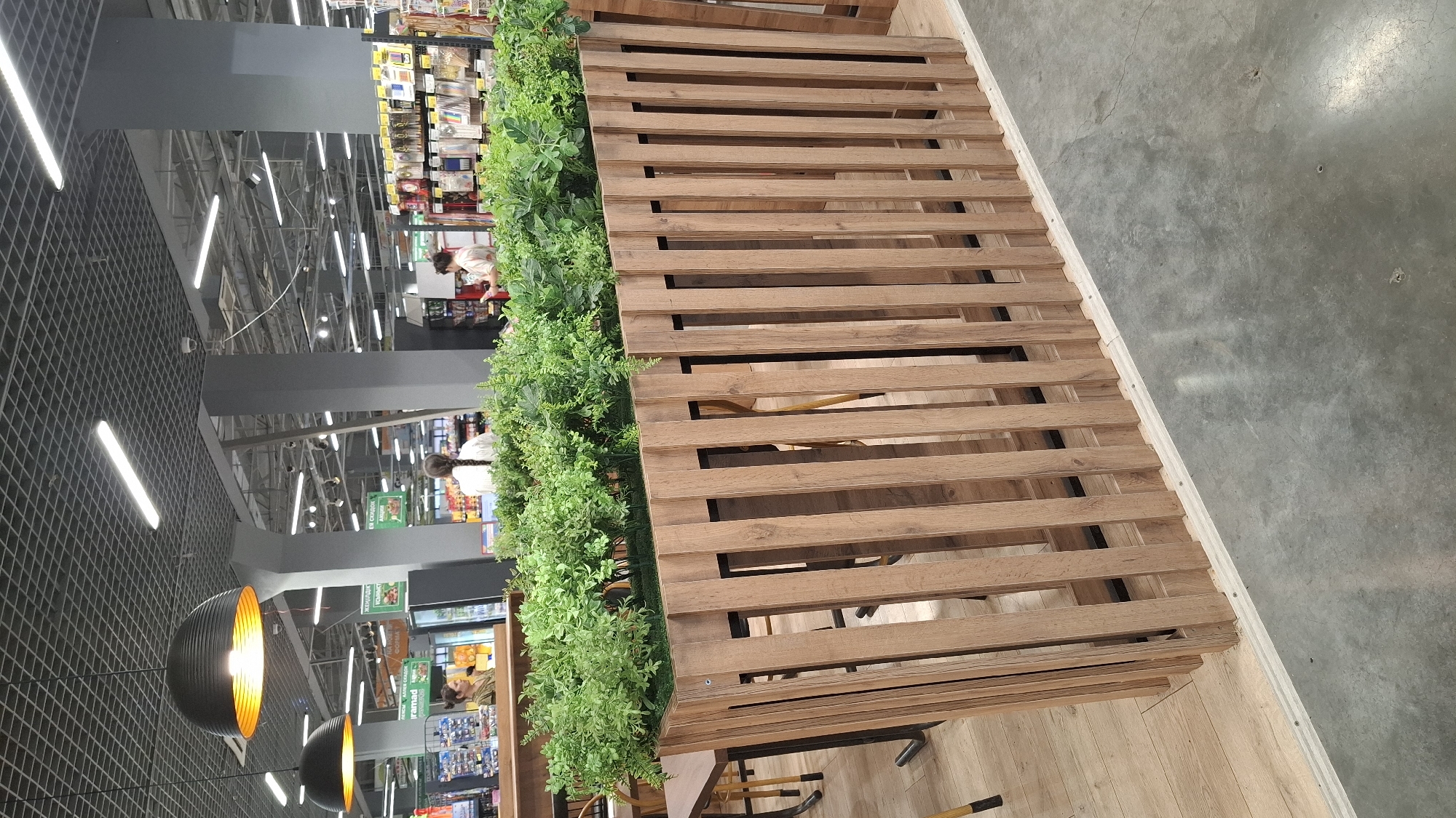 The image shows an indoor scene, likely a supermarket or a large retail store, in Shymkent, Kazakhstan. In the foreground, a wooden slatted partition with artificial greenery on top separates a seating area from the main aisle. The seating area has wooden tables and chairs, with a light wooden floor. Beyond the partition, the store's interior is visible, featuring rows of shelves stocked with merchandise. Several ceiling lamps, including large black pendant lights with orange interiors, illuminate the space. In the background, individuals are seen browsing or working. The overall environment appears to be a commercial setting.