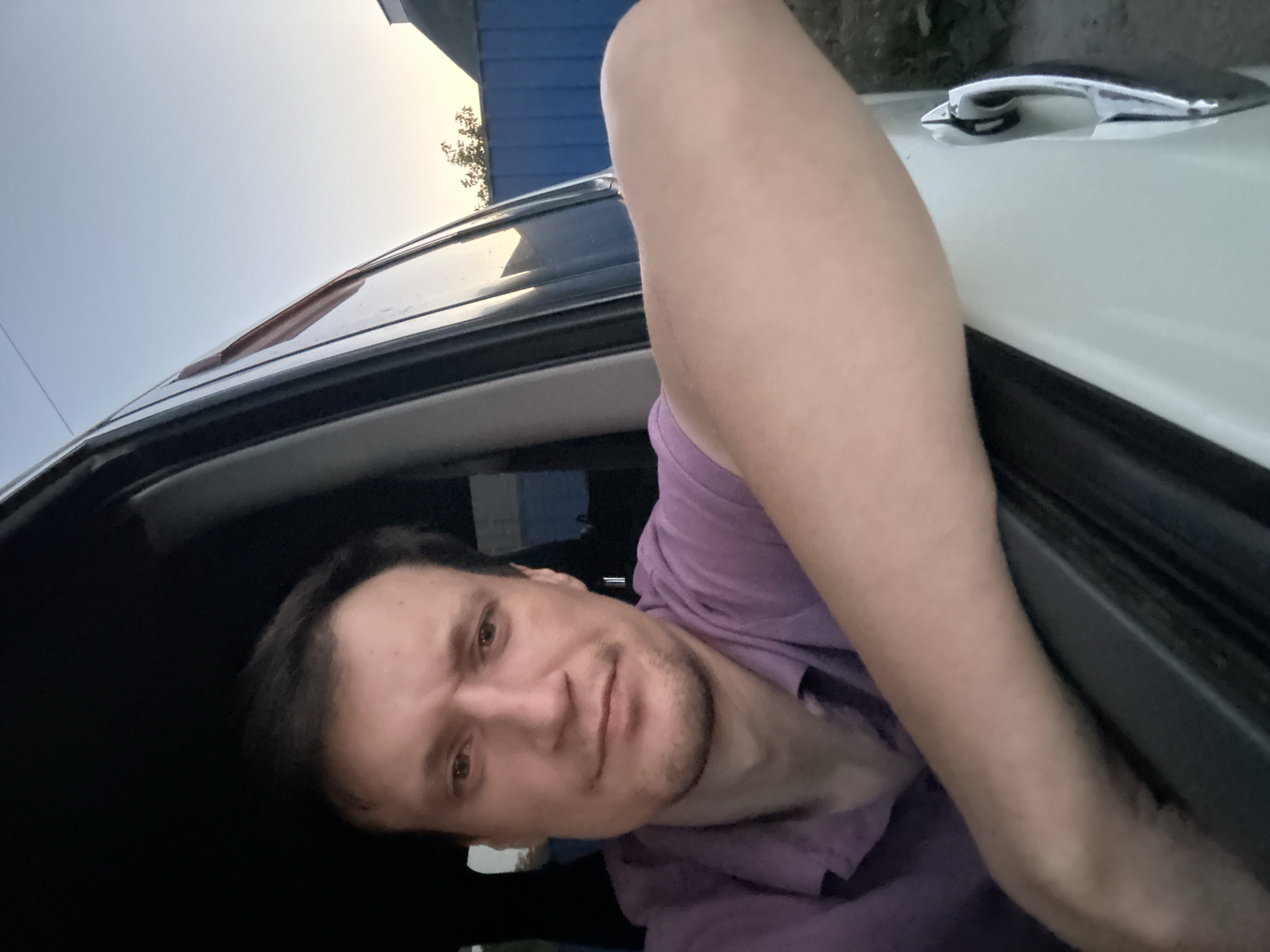 A man is leaning out of the driver's side window of a car. He is wearing a purple polo shirt and looking towards the camera with a slight frown. His right arm is resting on the car door. The car is white. In the background, a blue fence and some trees are visible under a pale sky. The image appears to have been taken outdoors in a rural or suburban setting, possibly in the city of Shatava, Ukraine.